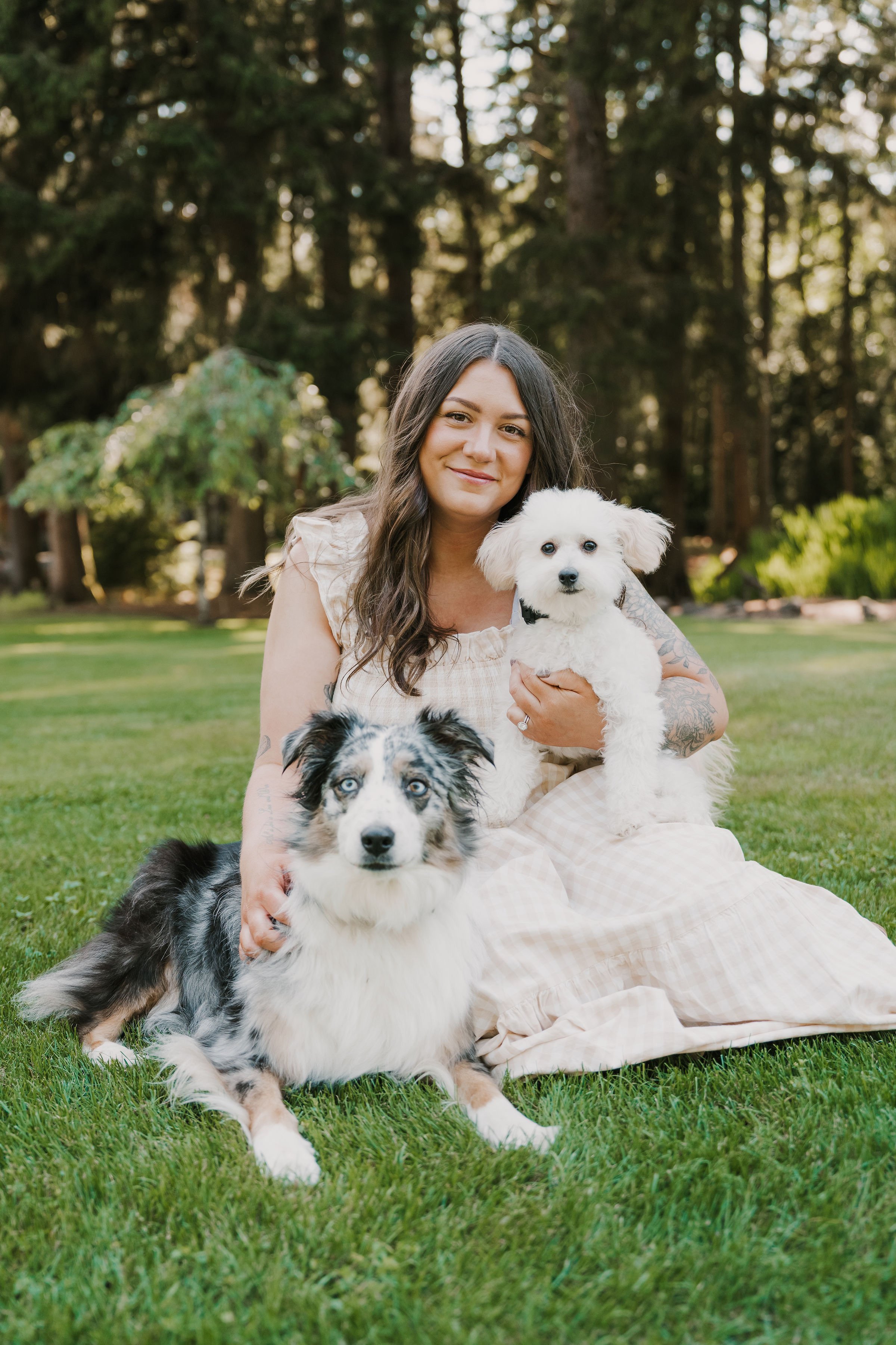 A woman with long dark hair and tattoos on her arm sitting on grass in a park, holding a small white dog. Two other dogs are also near her, one with black and white fur and blue eyes, and the other with a black and white merle coat. Tall trees are in the background.