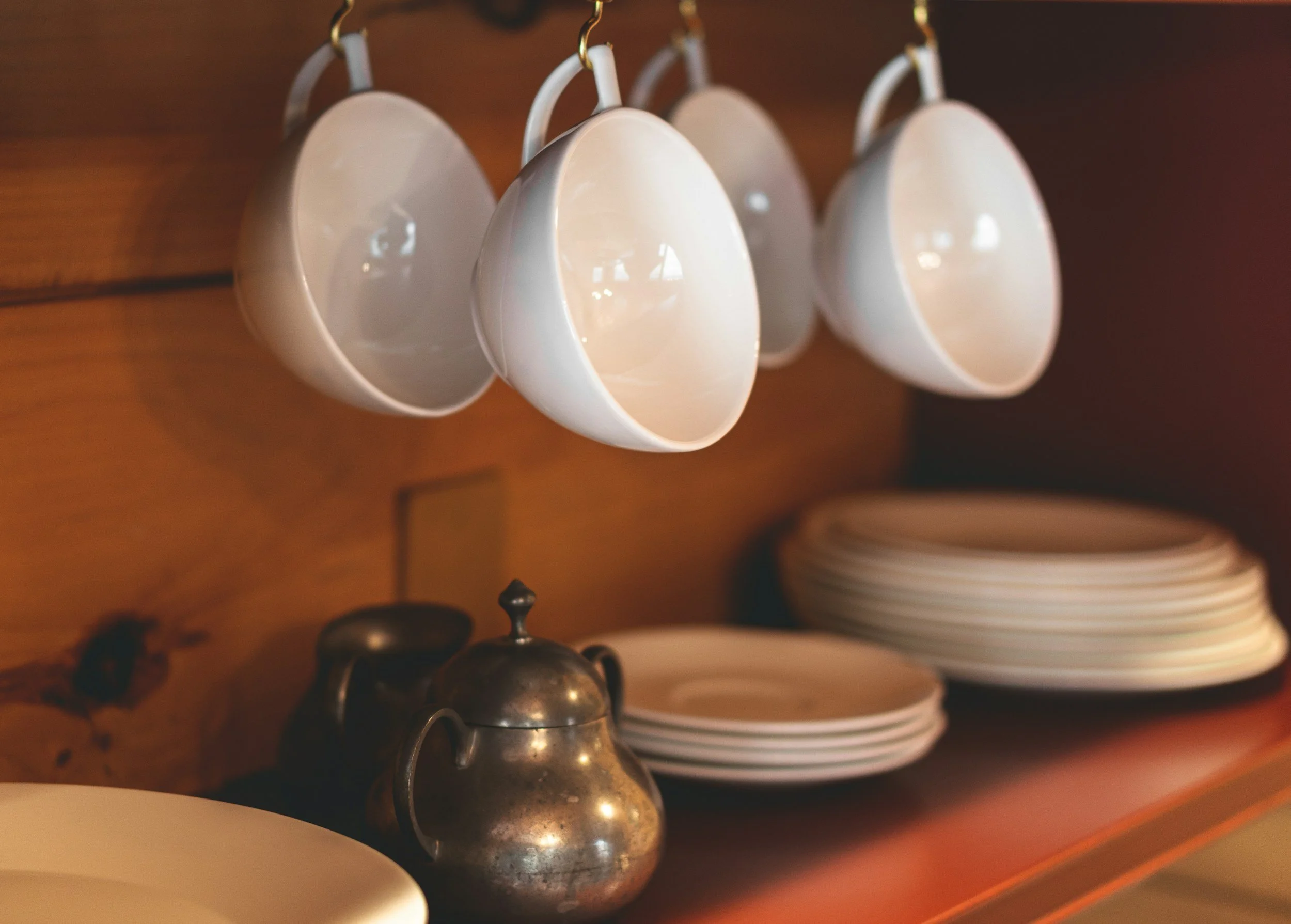 A metal bell and several white ceramic cups hanging on hooks on a wooden wall, with stacked white plates visible in the background.