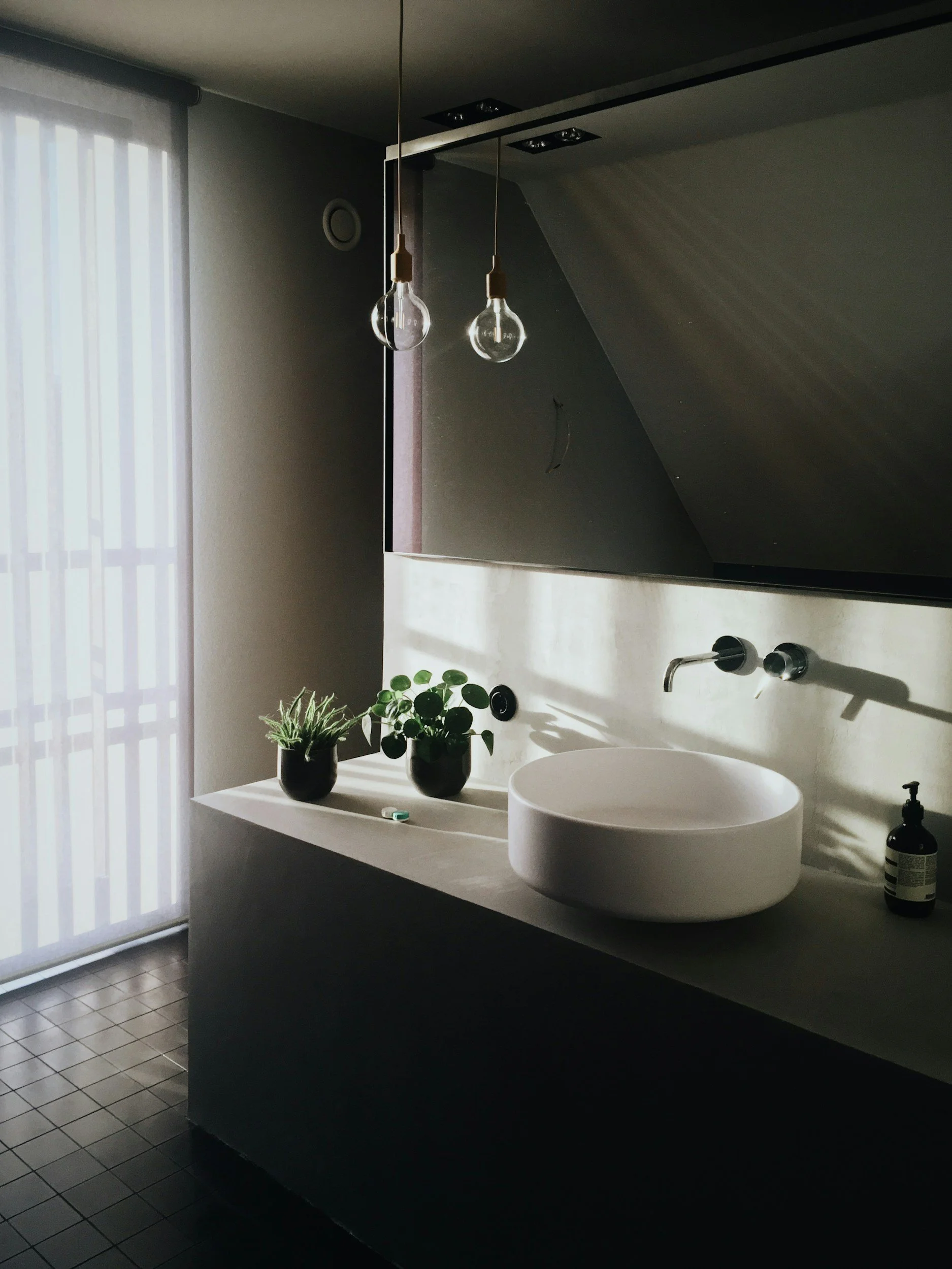 A modern bathroom with a white vessel sink on a black countertop, decorated with potted plants, and a large mirror reflecting two bare light bulbs hanging from the ceiling. Sunlight filters through sheer curtains on a window or door to the left.