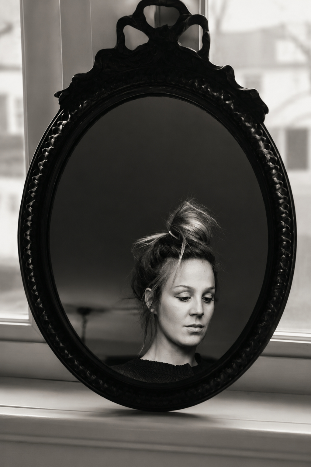 A black and white photo of a woman looking into an ornate oval mirror with a dark frame, her hair styled in a messy bun.