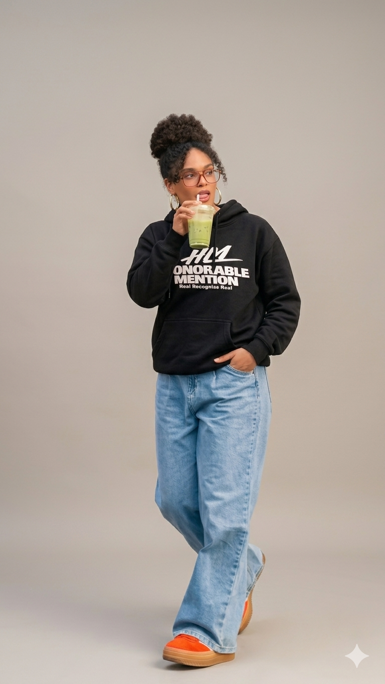 A woman with curly hair in a high puff, wearing glasses, a black hoodie with white text, light blue jeans, and orange shoes, holding a green drink in a clear cup and standing against a plain light gray background.