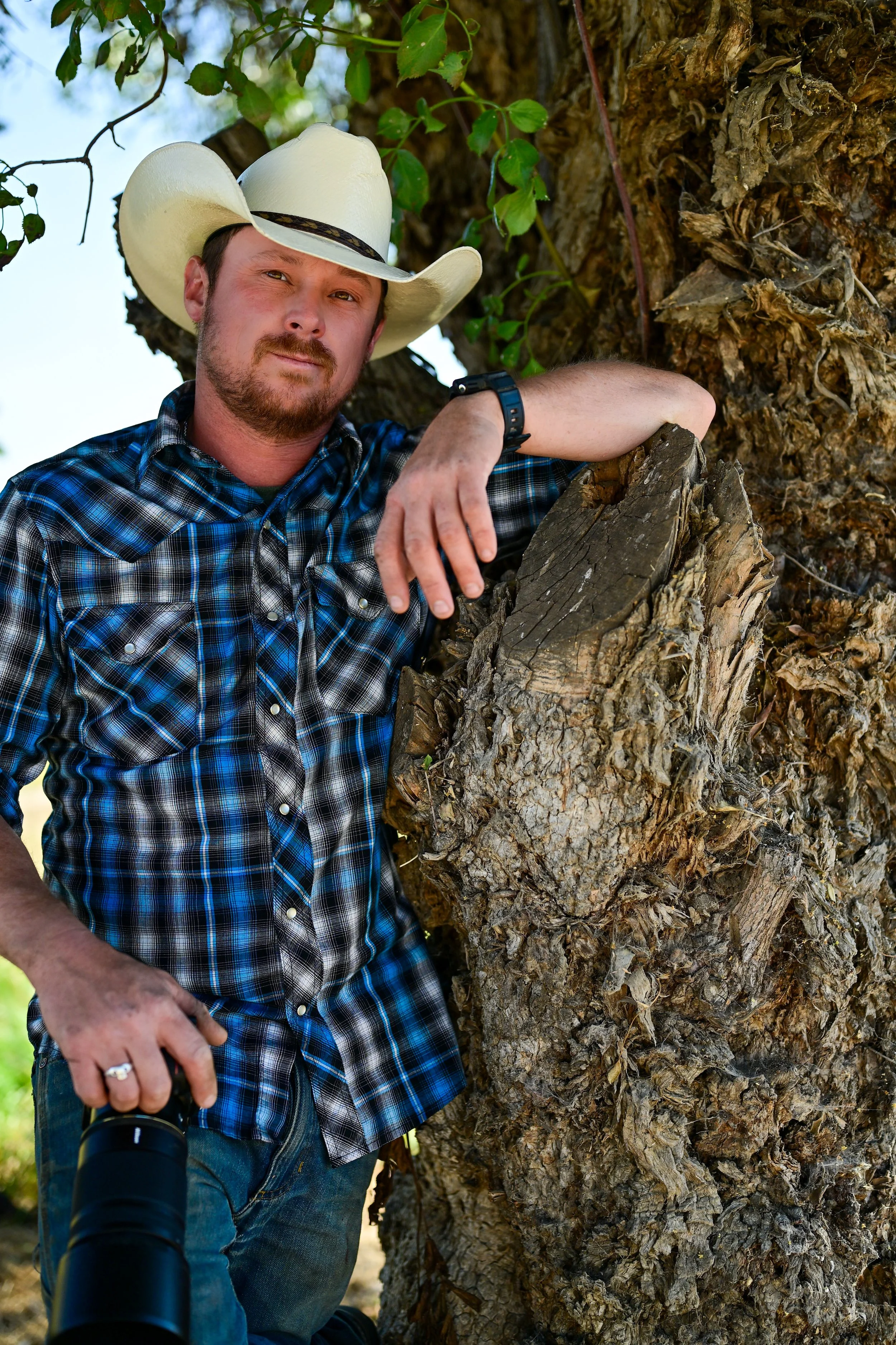 A man in a plaid shirt and cowboy hat leaning on a tree trunk, holding a camera with a telephoto lens.
