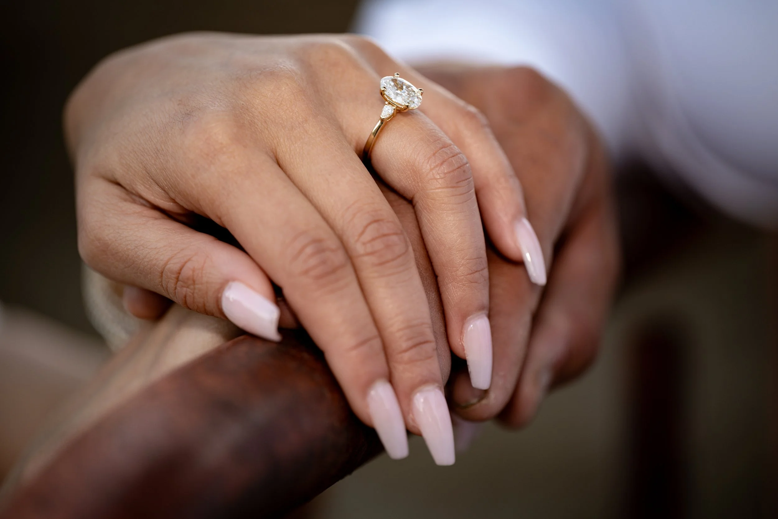 Close-up of a person’s hand wearing a gold ring with a large oval-cut diamond. The background is blurred, focusing on the ring and hand.