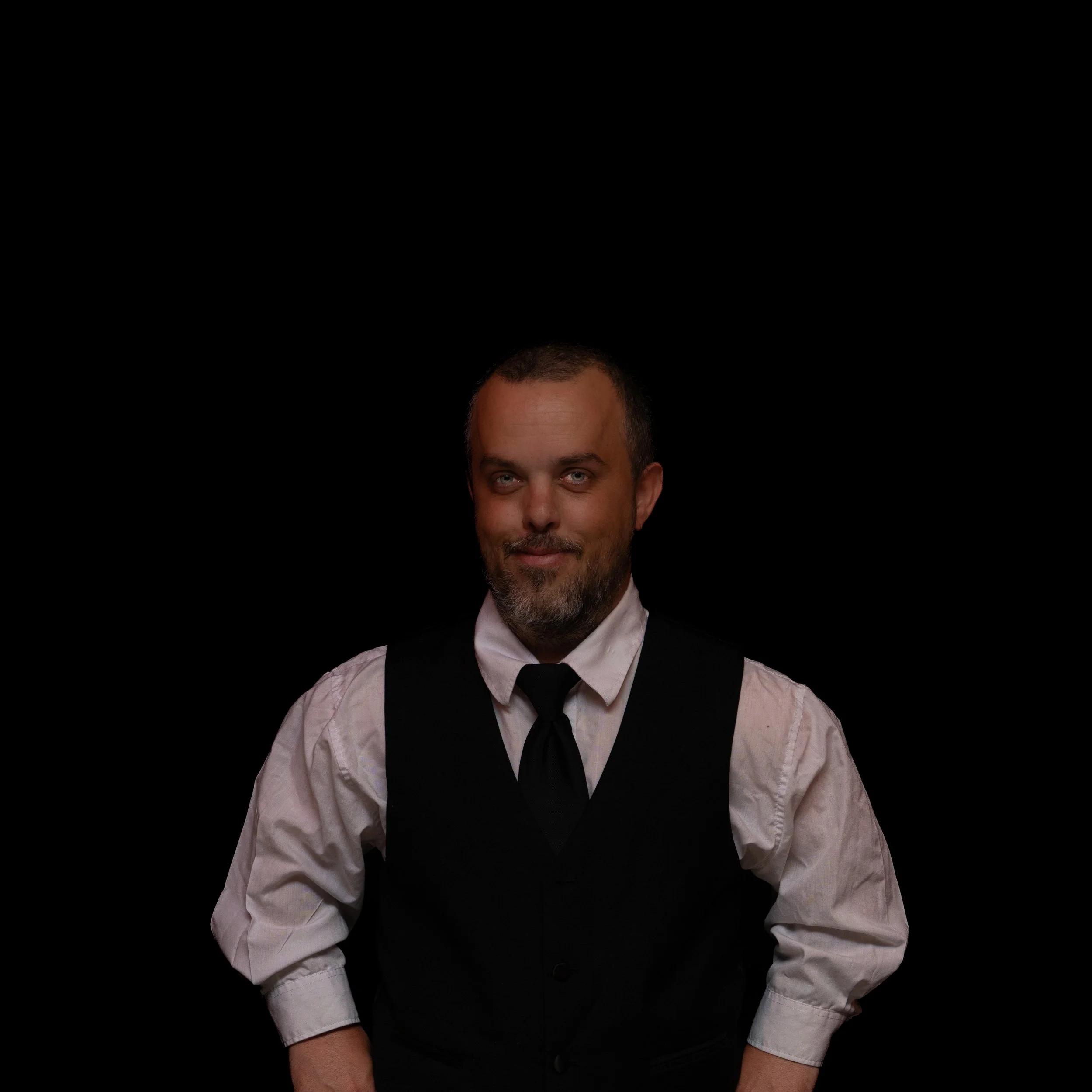 Portrait of a man with short hair and a beard, wearing a white shirt and a black vest, against a black background.