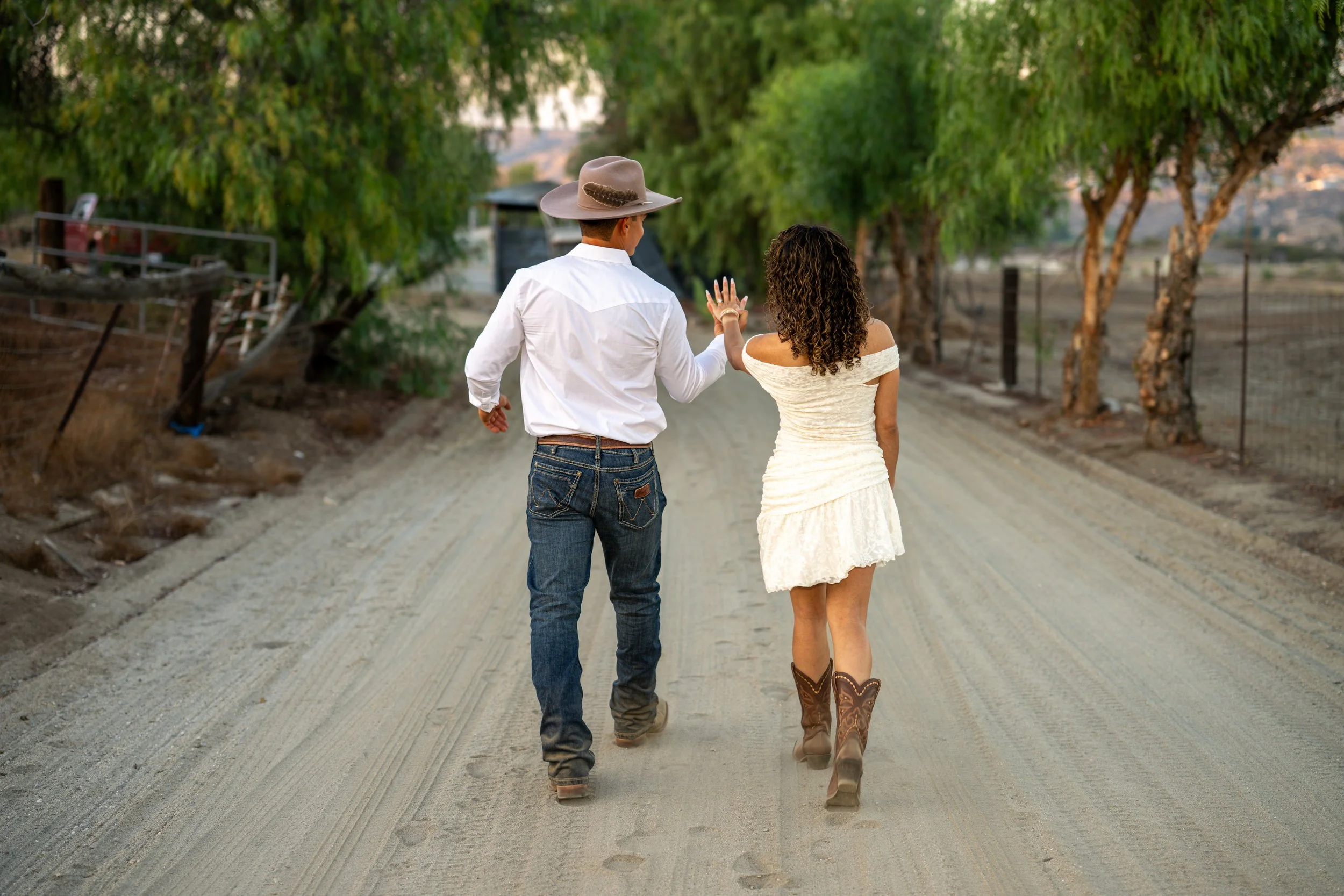 A man and woman walking on a dirt road in a rural area, surrounded by trees. The man is wearing a cowboy hat, white shirt, jeans, and boots, while the woman is wearing an off-the-shoulder white dress and cowboy boots. They appear to be engaged in conversation and are seen from behind.