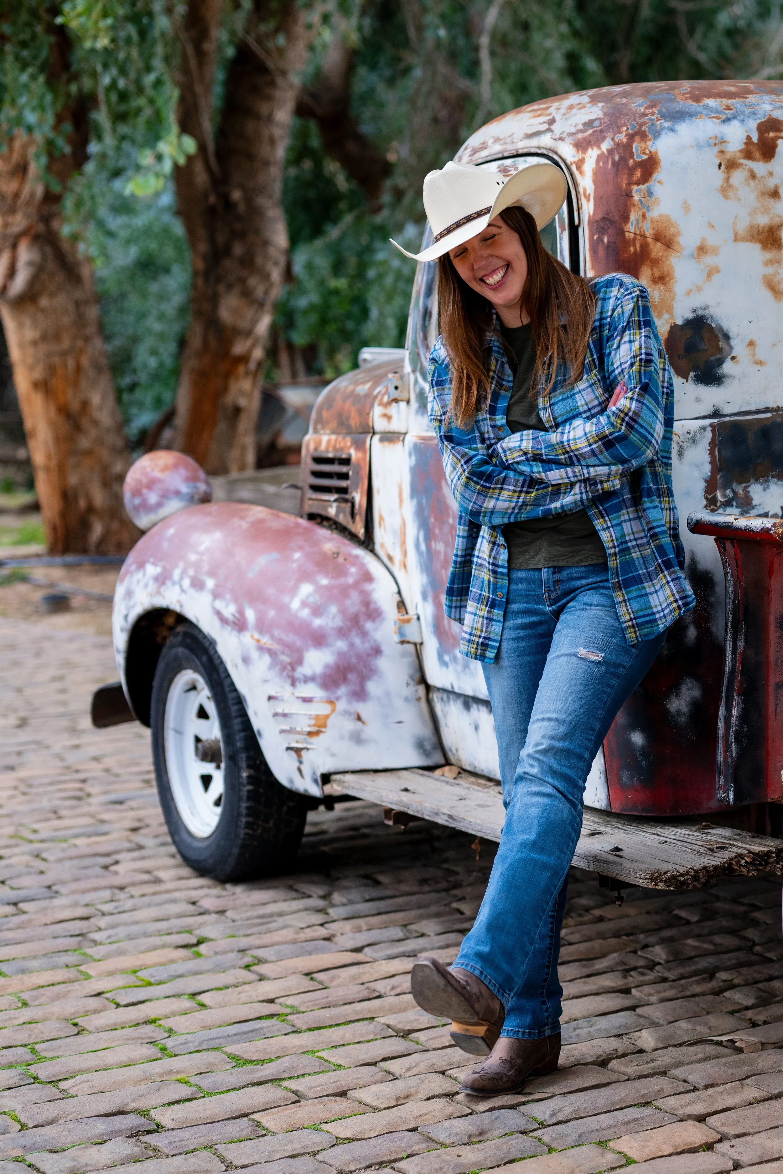 A woman wearing a cowboy hat and plaid shirt leaning against a rusty, vintage pickup truck on cobblestone ground, smiling with her eyes closed.