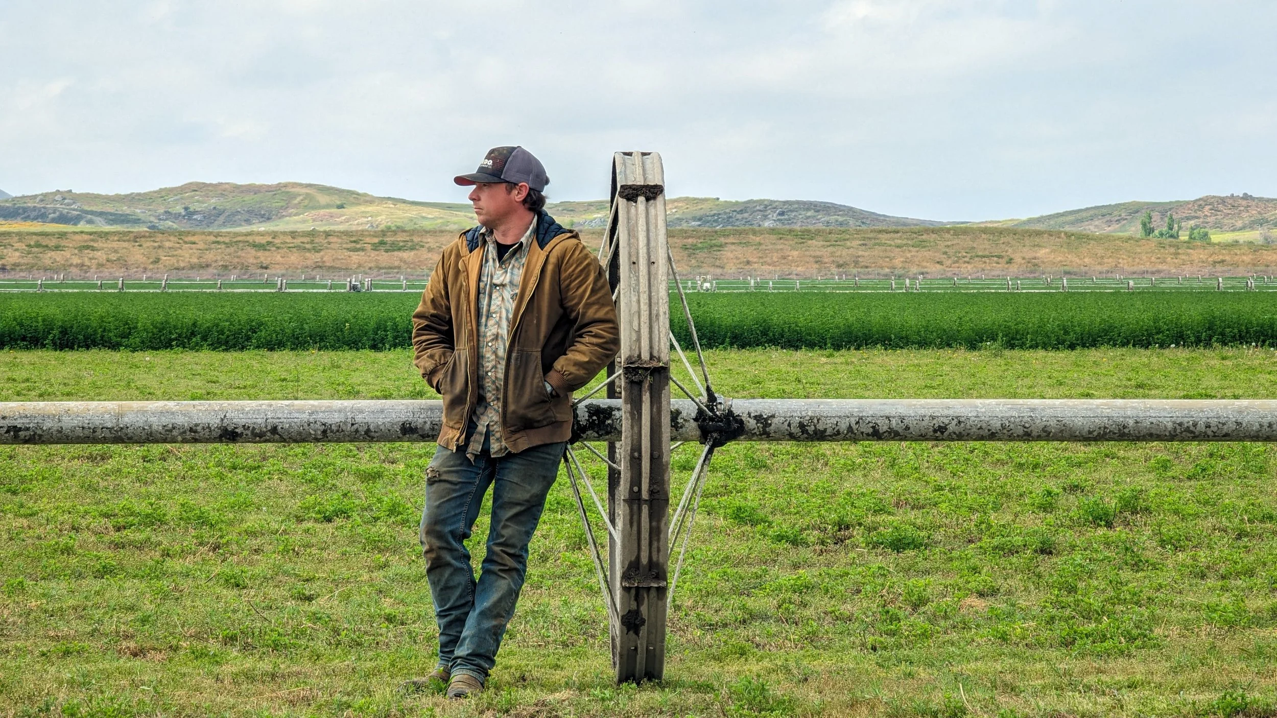 A man in a brown jacket and baseball cap leaning against a metal pole in a rural field with green grass and hills in the background.