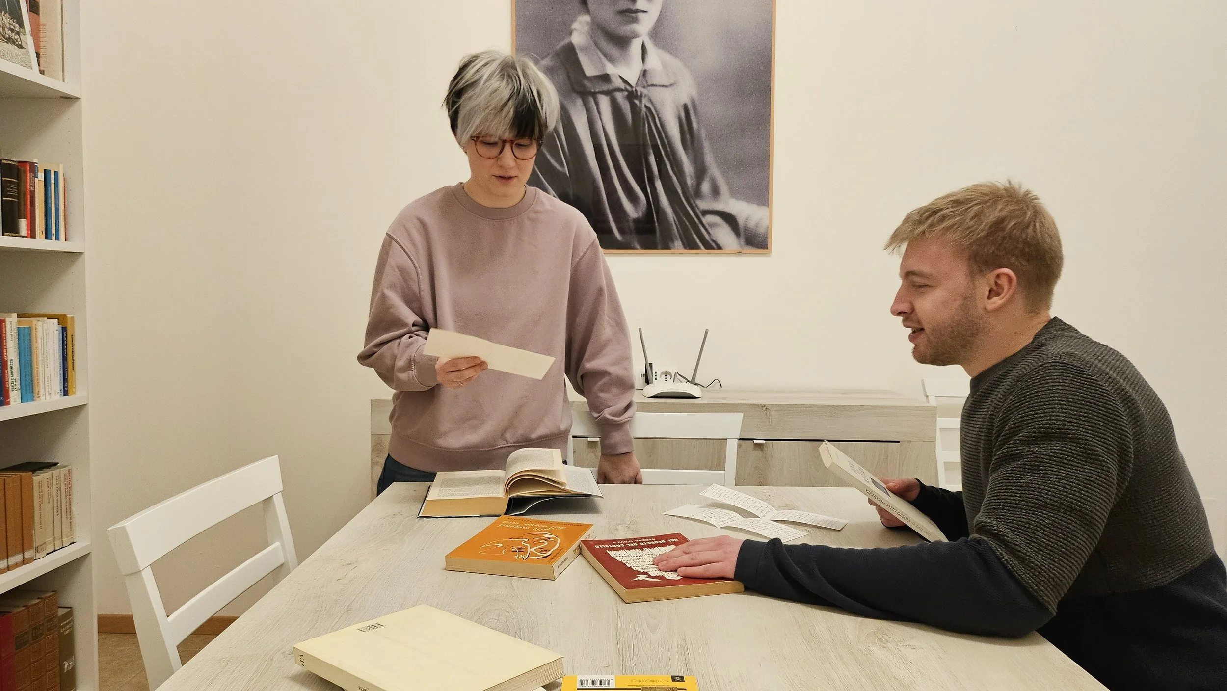 Two people are seated at a table with books and papers, while another person stands, looking at papers. The setting appears to be a study or library room with a bookshelf on the left and a black-and-white portrait on the wall.