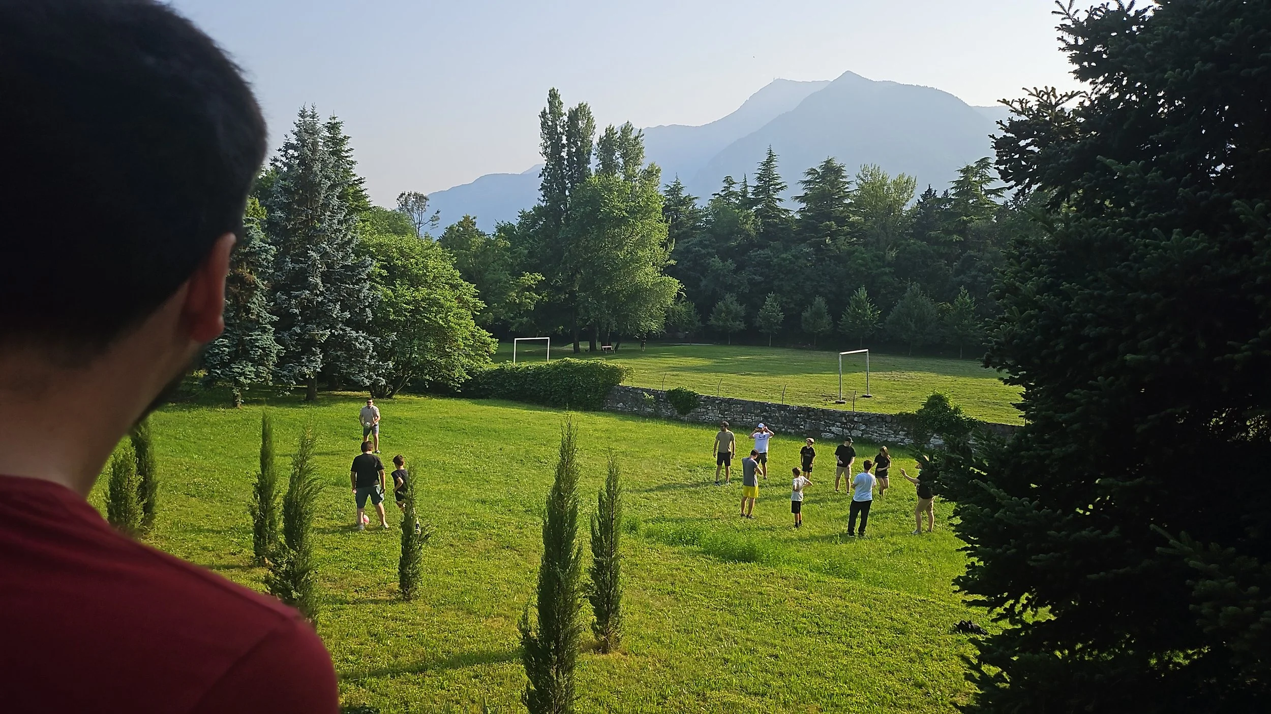 A person with short hair and a red shirt in the foreground looking at a group of people in the garden of Le Laste outdoor field surrounded by trees and mountains in the background.