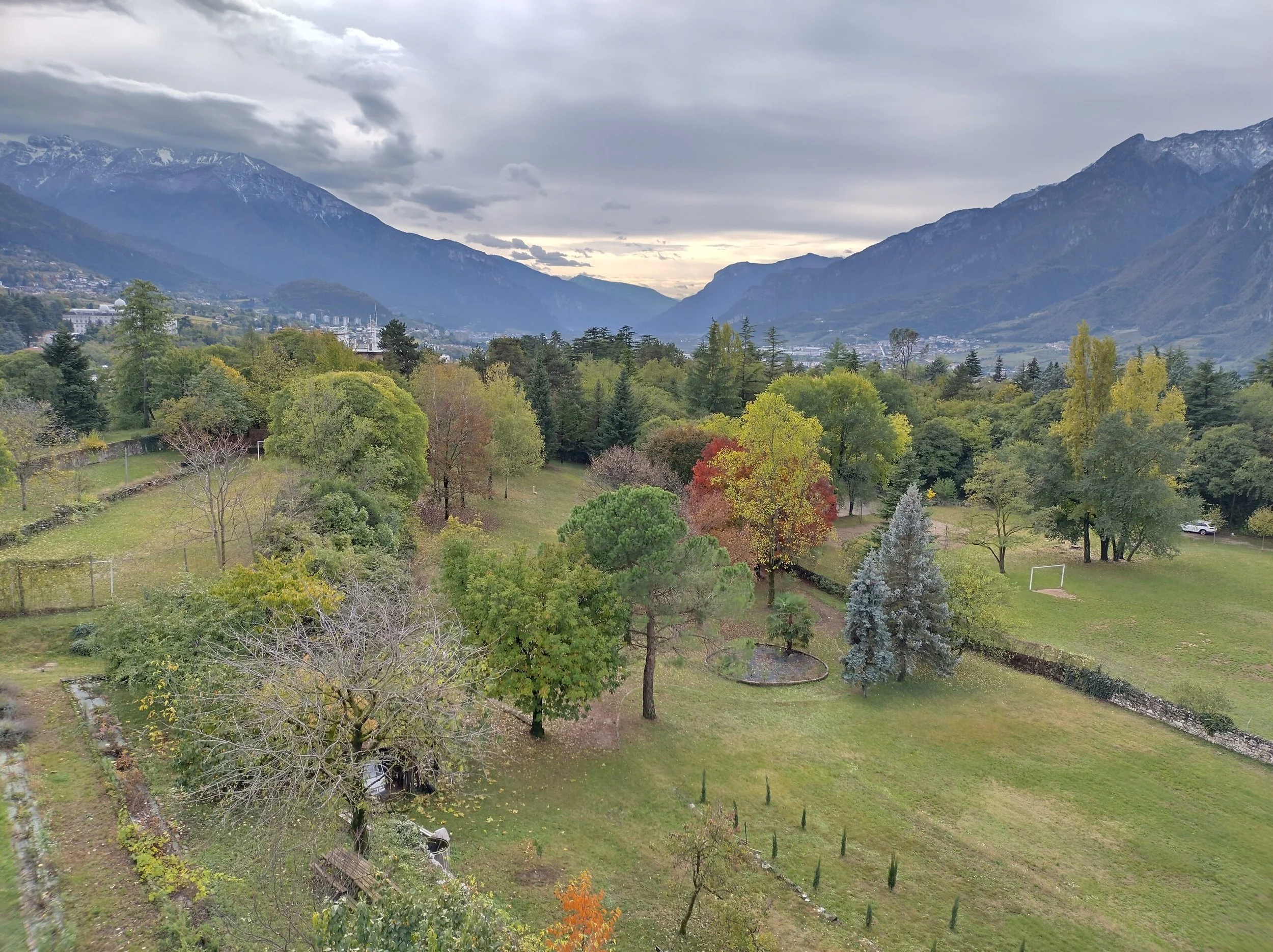A scenic view of a lush green park Le Laste with various trees, beyond which is a small town, surrounded by tall mountains under a cloudy sky.