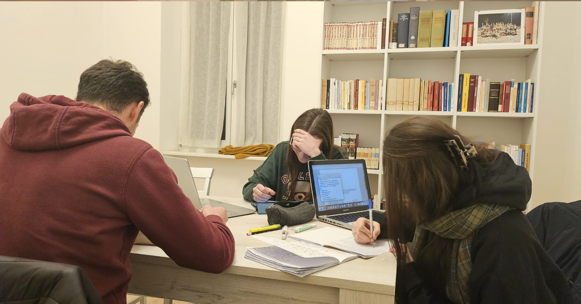 Three people sit at a table in a room with a bookshelf filled with books in the background. They are working with laptops, notebooks, and writing utensils, appearing focused and stressed, with one person holding their forehead.