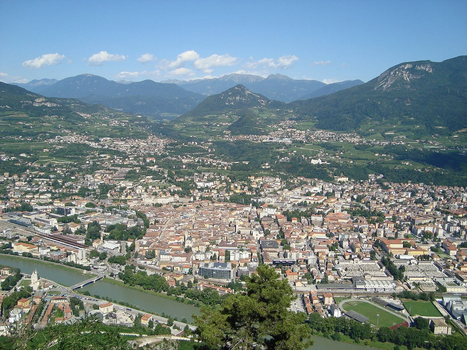 A panoramic view of a European city with a river running through it, surrounded by green mountains under a blue sky with scattered clouds.