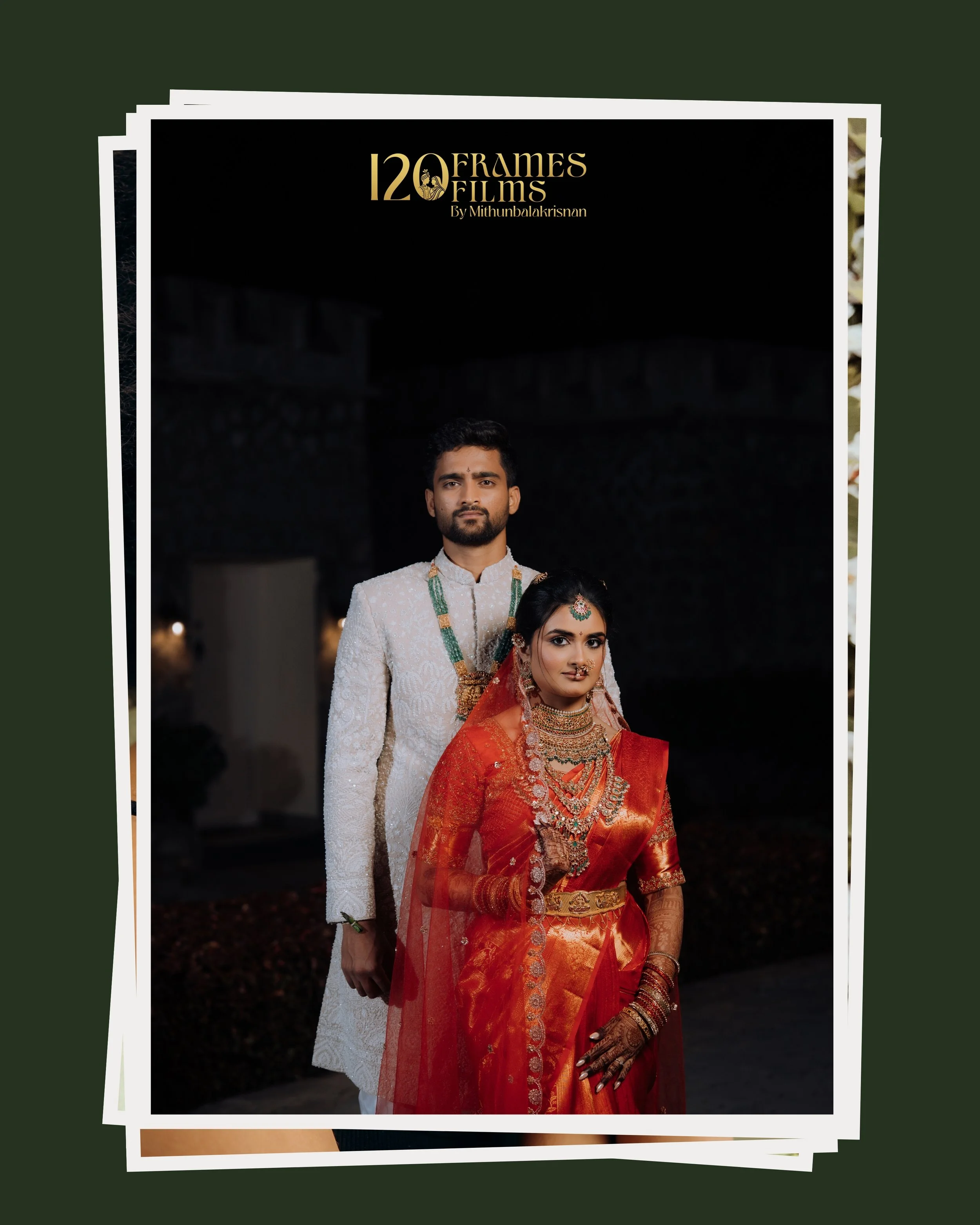 A couple dressed in traditional Indian wedding attire standing outdoors at night. The man is in a cream-colored sherwani with green jewelry, and the woman is in a red saree with extensive gold jewelry and henna on her hands.