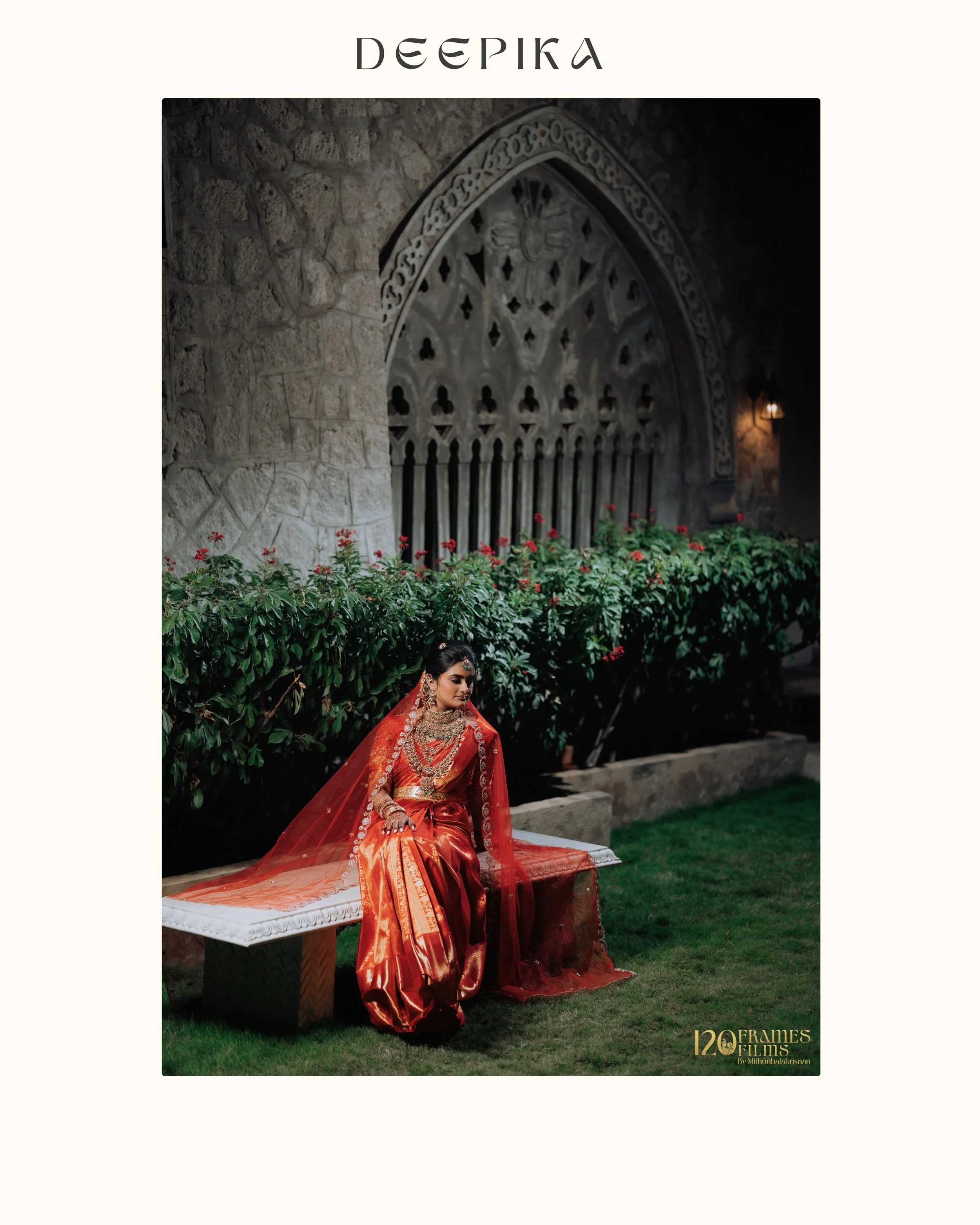 A woman dressed in traditional red and gold Indian attire sits on a white ornate bench next to a lush green bush with red flowers, in an outdoor garden setting with stone wall and decorative window in the background.
