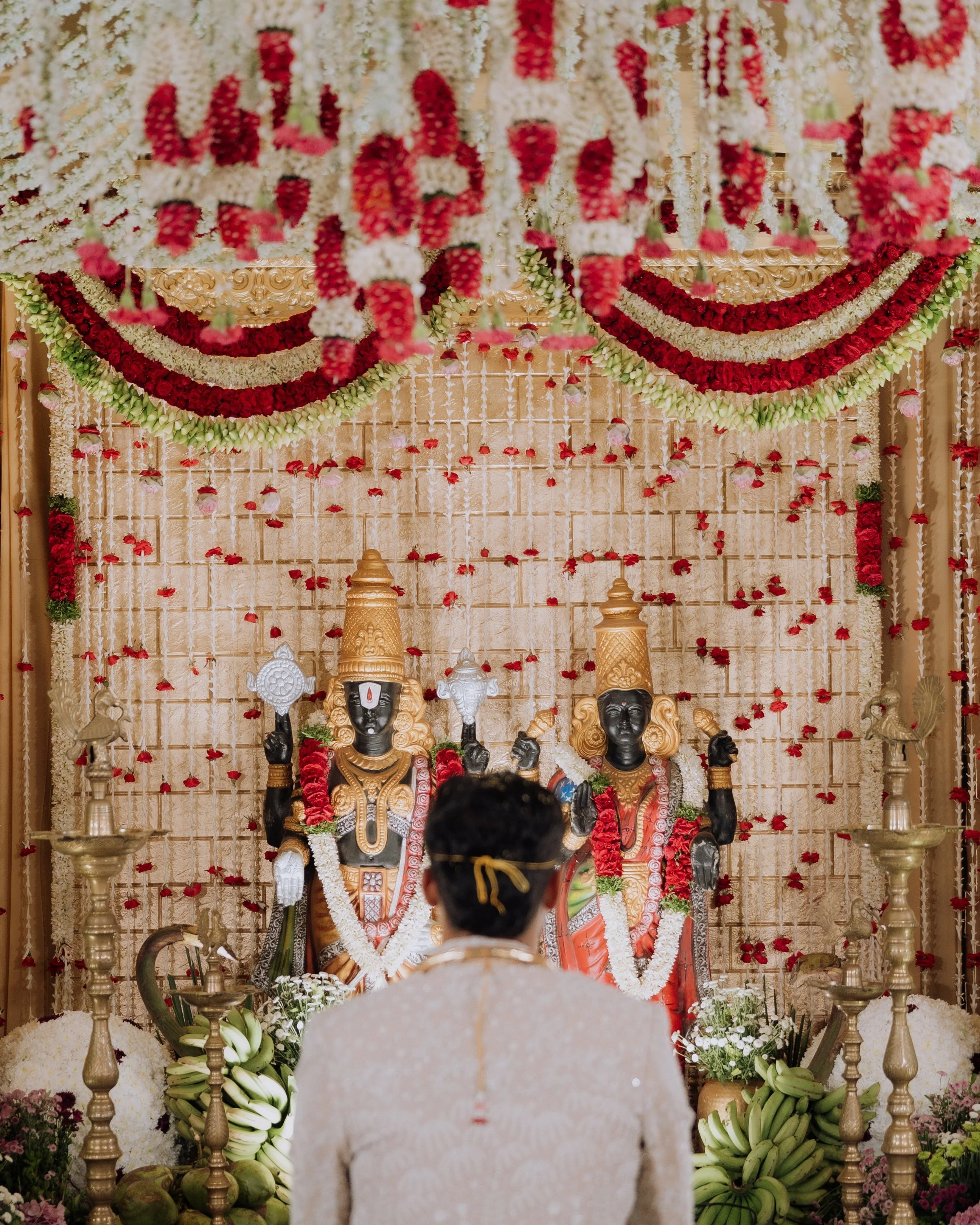 A traditional Indian wedding altar decorated with flowers and idols of deities, with a bride and groom in traditional attire seated in front.