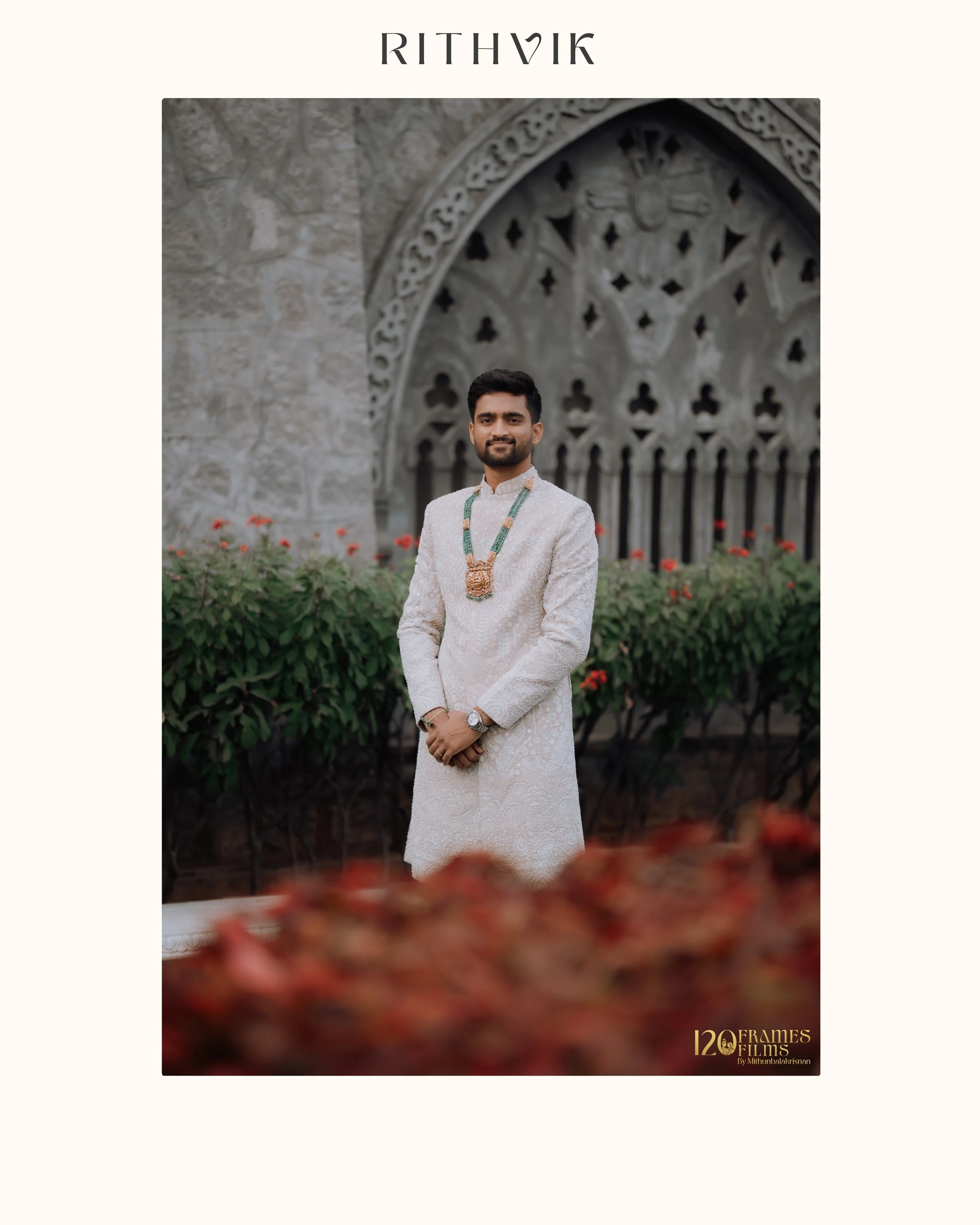 Man in traditional cream-colored ethnic attire with green beaded necklace standing outdoors in front of an ornate stone archway, with flowers and greenery around.
