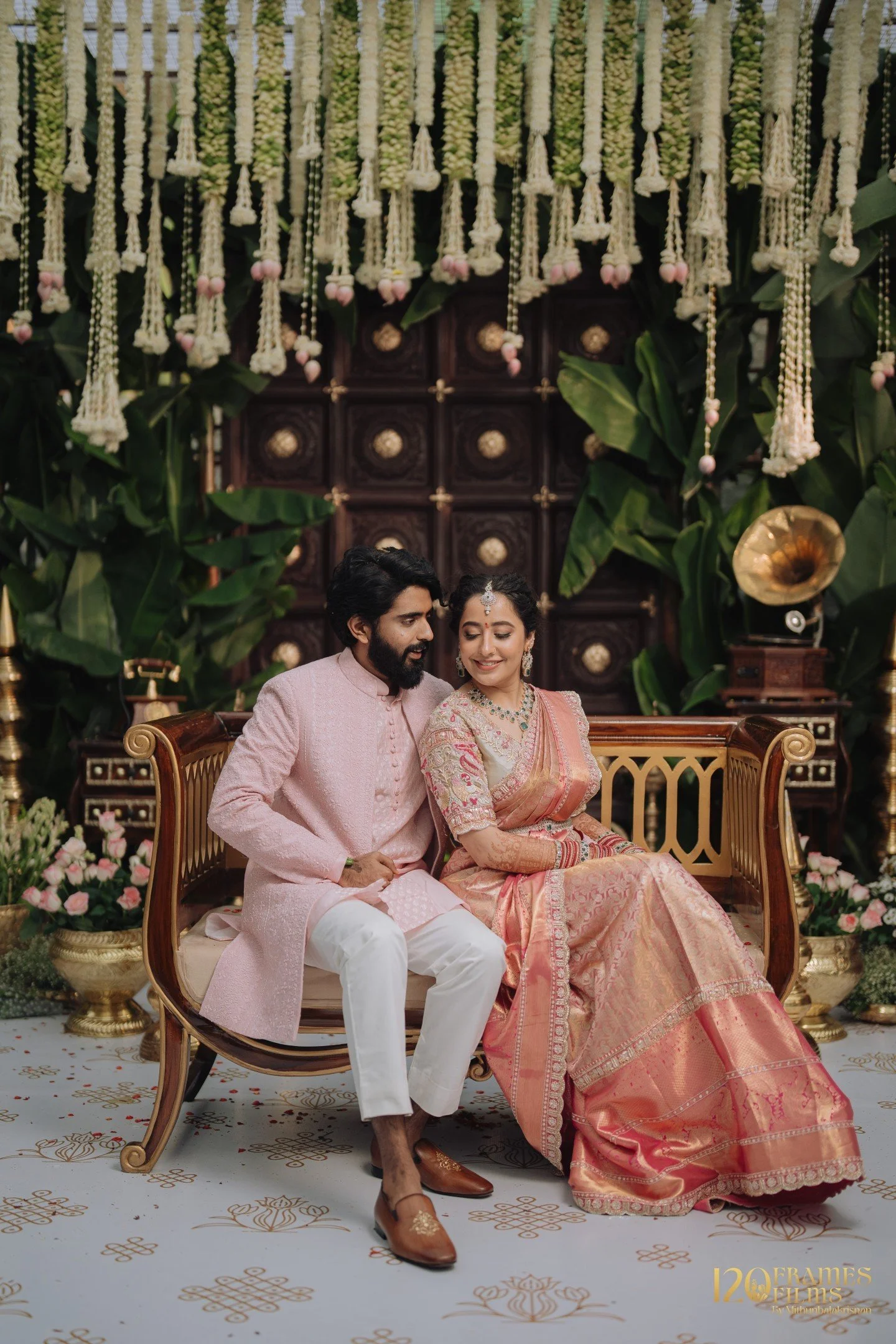 A couple dressed in traditional Indian wedding attire sitting on a decorated wooden bench, surrounded by floral arrangements and lush greenery in the background.