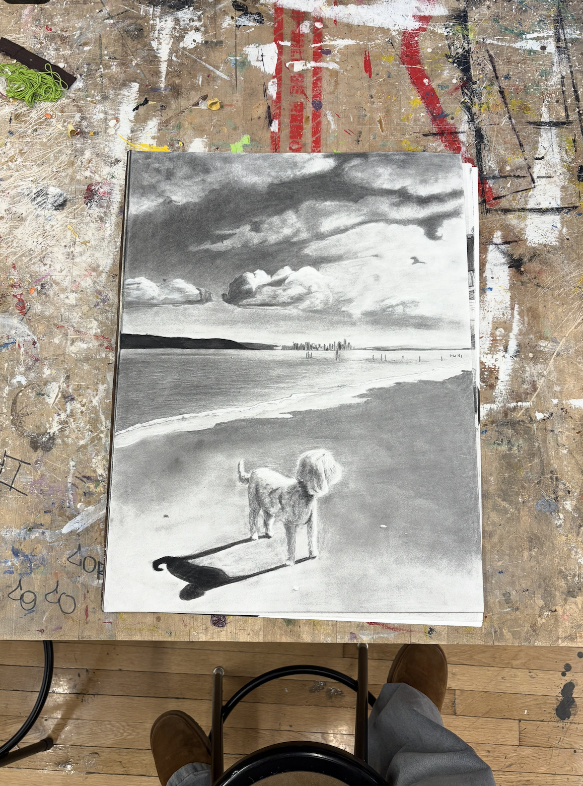Black and white painting of a dog standing on a beach with a city skyline in the distance, dramatic cloudy sky, seen from above on a cluttered art table.