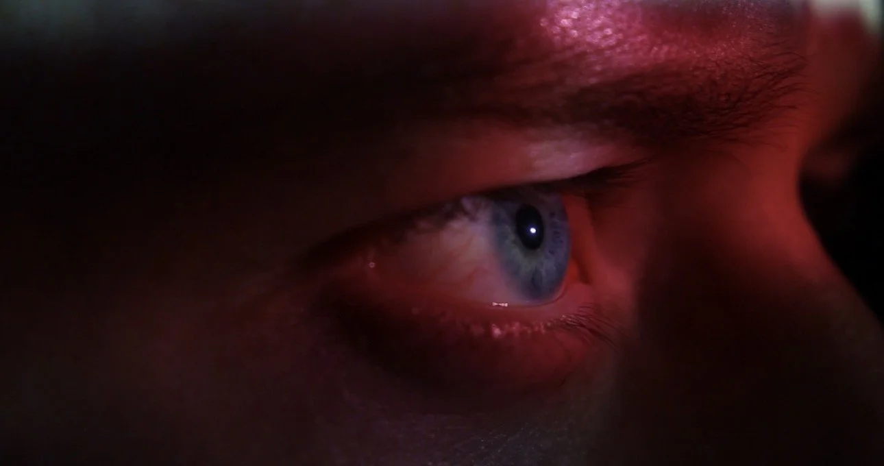 Close-up of a person's eye with a blue iris, partially covered by eyelid, in a dimly lit environment with red lighting.