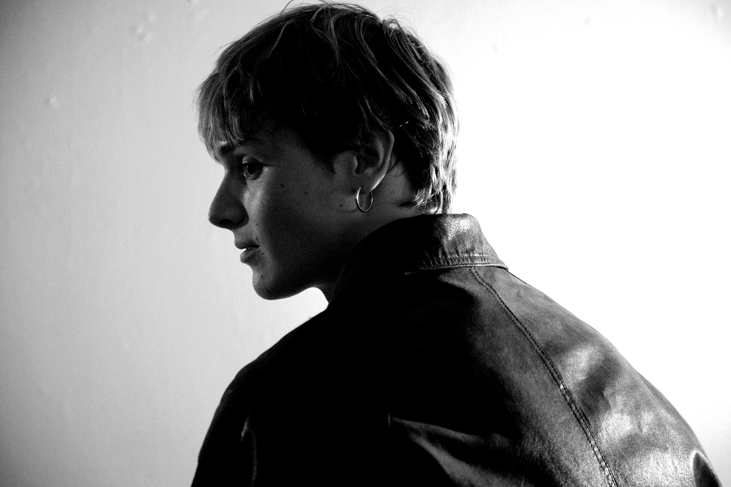 A black-and-white side profile of a young person with short, tousled hair, wearing a hoop earring and a leather jacket.