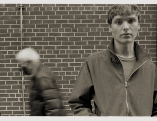 A young man with straight brown hair and a neutral expression standing against a brick wall, with an older person wearing a puffy jacket walking by in the background.