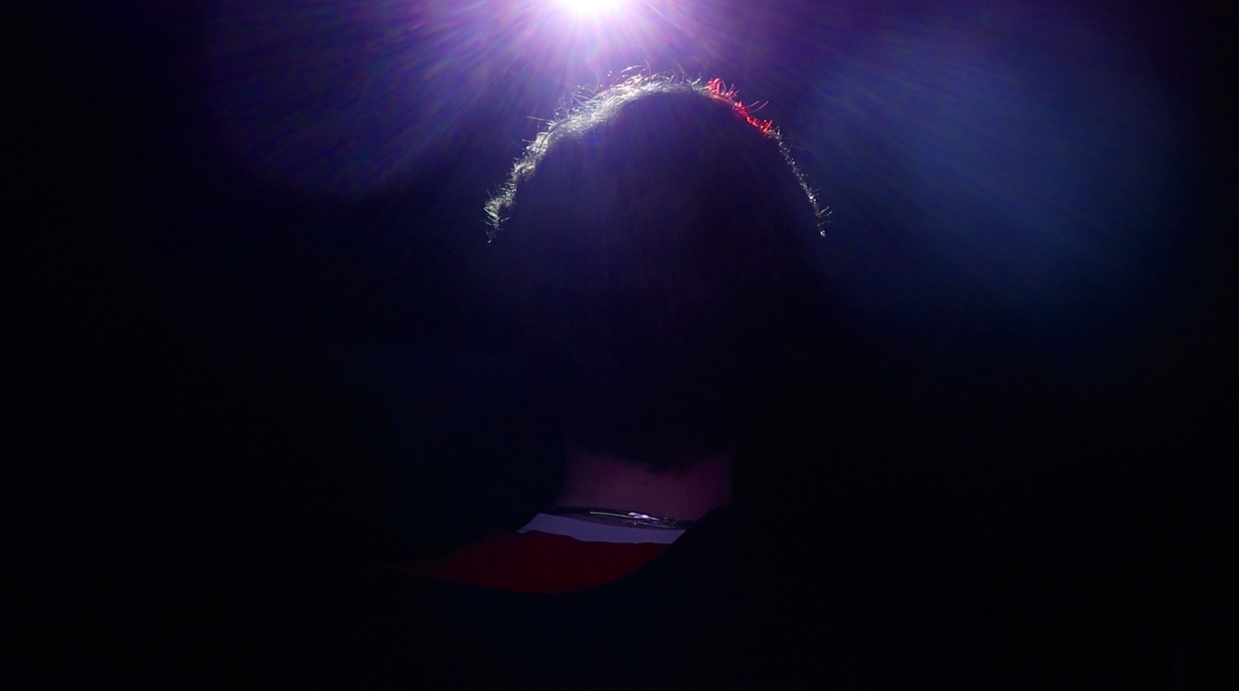 Person with curly hair sitting in front of a bright light shining down from above into their head, creating a glow and lens flare effect.
