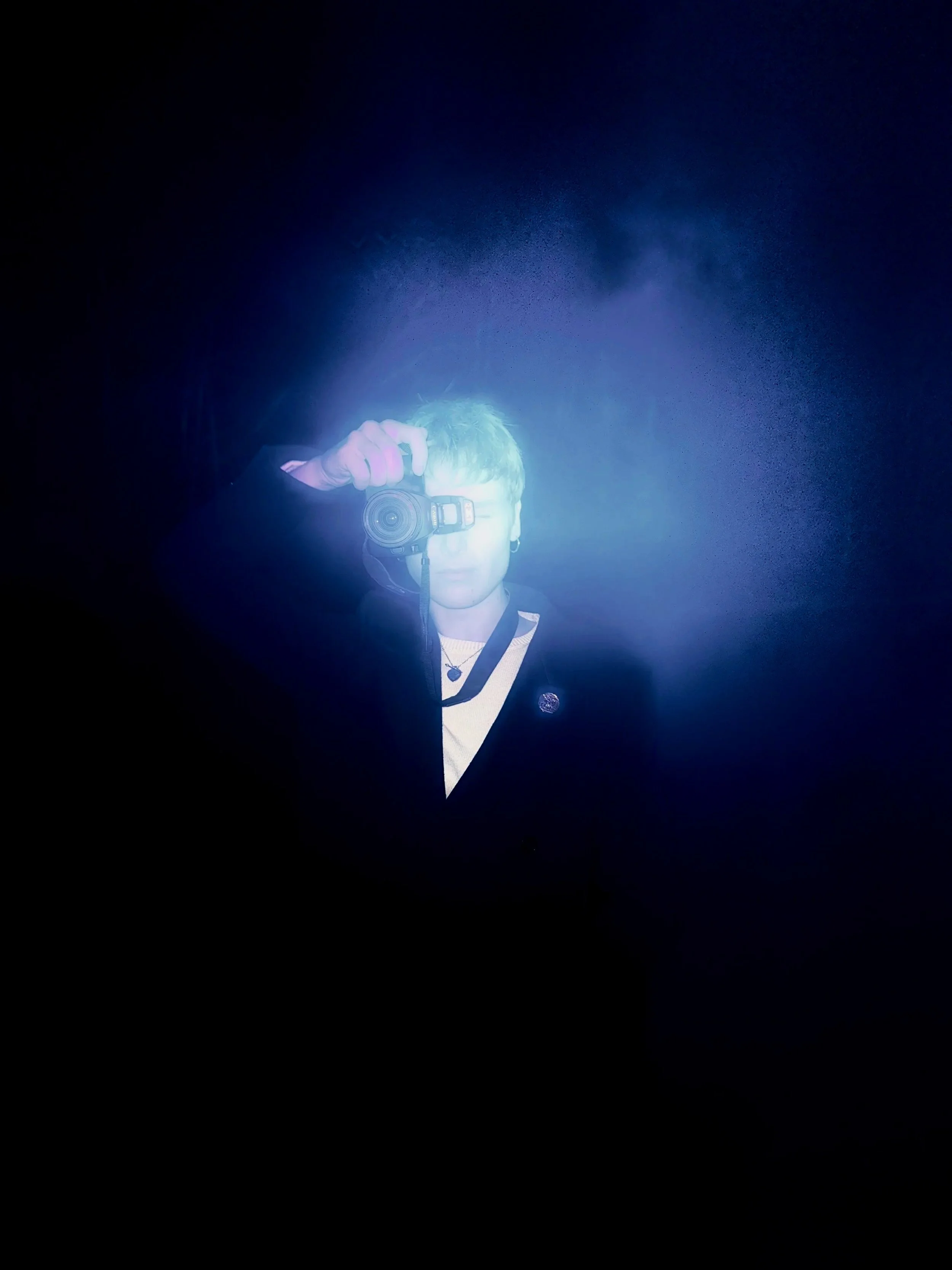 Person taking a selfie with a camera in front of a dark background, with blue lighting reflecting on their face.