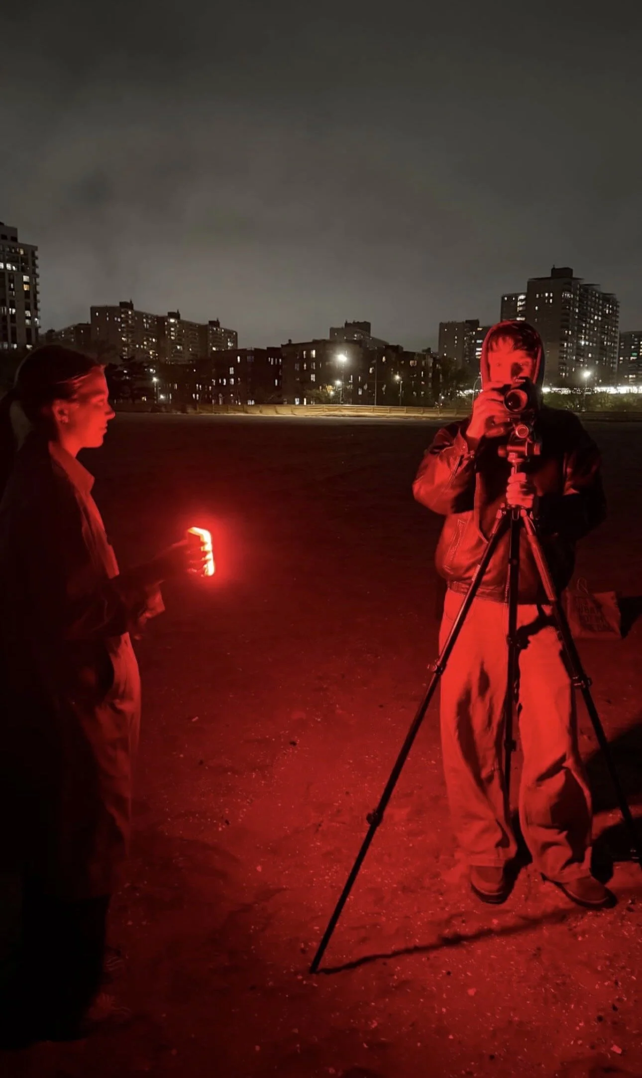 Two people standing outdoors at night among city buildings, with one person holding a red light and the other taking a photograph with a camera on a tripod, illuminated by red and ambient city lights.