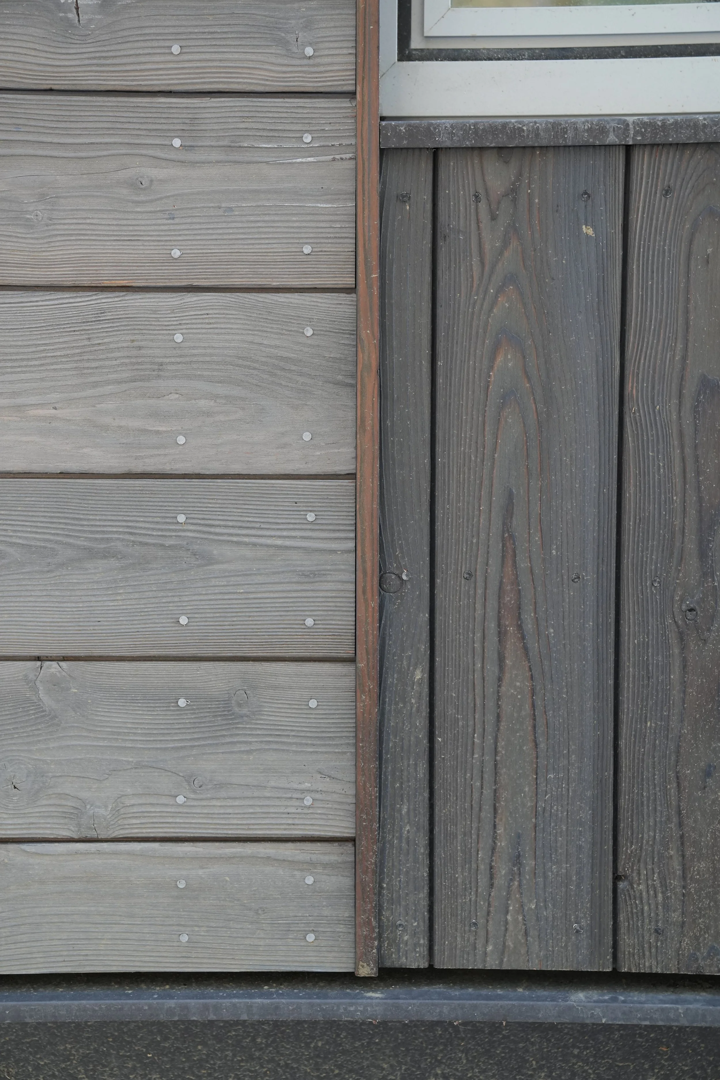 Close-up photo of a wooden exterior wall with horizontal wooden planks on the left and vertical wooden planks on the right, separated by a narrow vertical strip. The left side has lighter, weathered wood with visible nails, while the right side has d