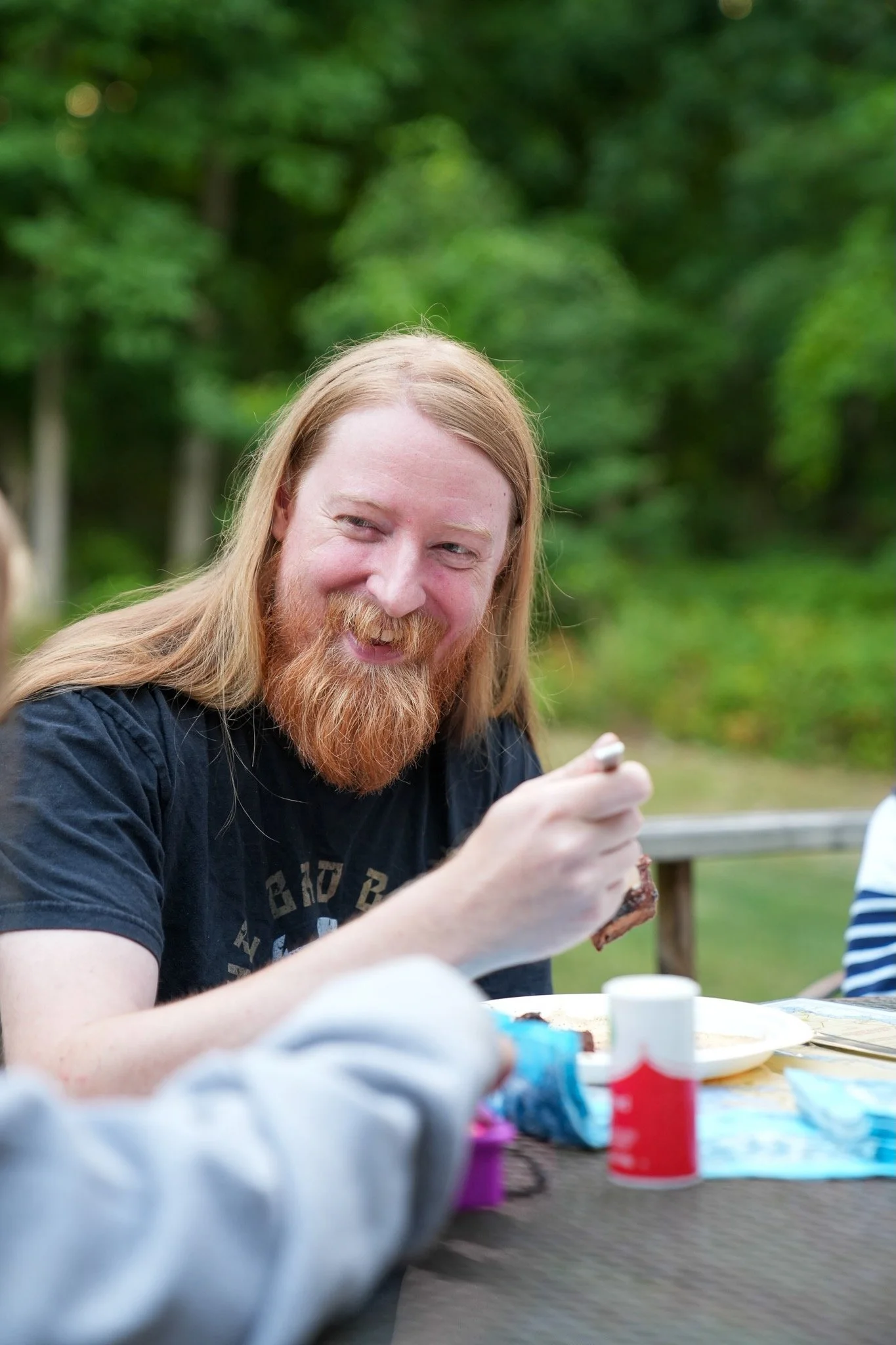 A man with long red hair and a beard smiling while sitting at a table outdoors, holding a piece of food. There are food items and bottles on the table. The background shows green trees.
