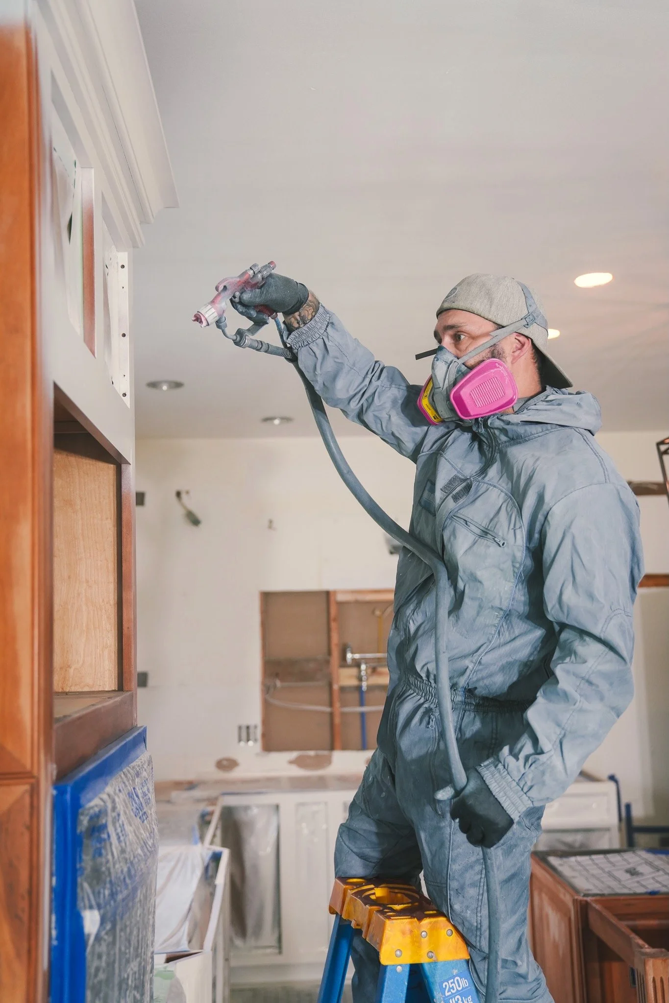 A man using a spray gun while painting or staining wooden cabinets or furniture inside a room. He is standing on a ladder, wearing protective gear including a mask and gloves.