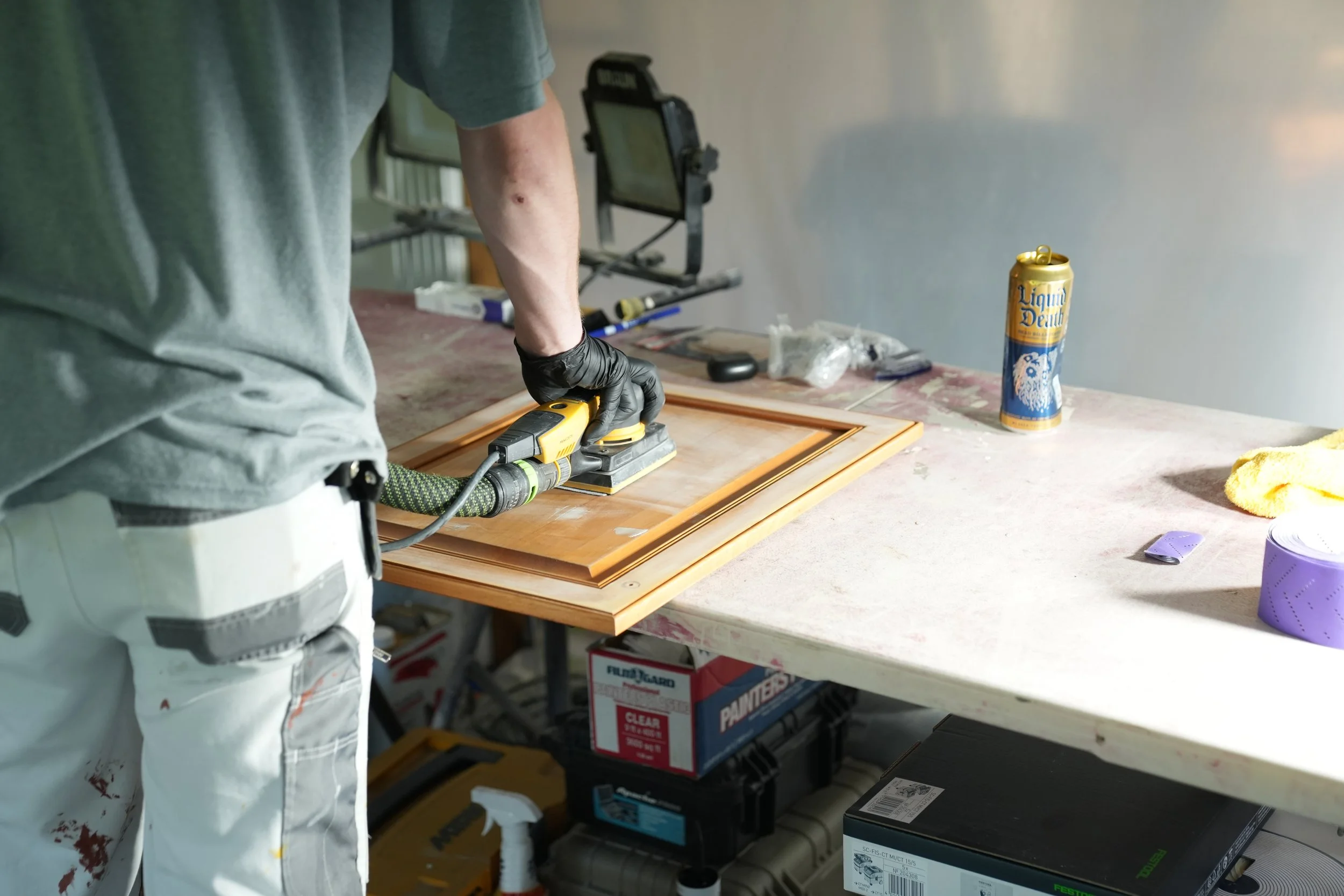 Person wearing gloves sanding a wooden picture frame in a workshop, with tools and supplies on the work surface and boxes underneath.
