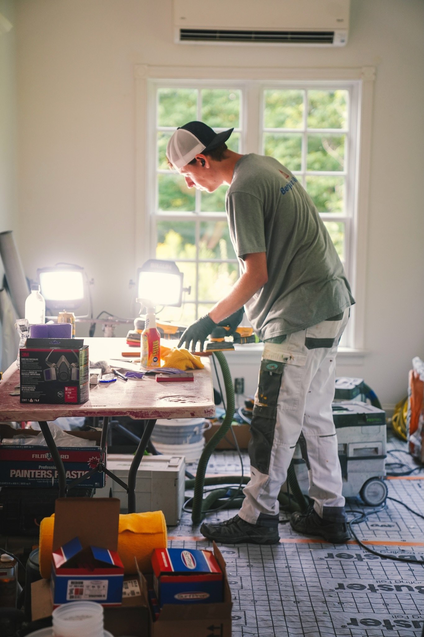 A man wearing work clothes, including a white cap, gloves, and safety shoes, is working in a room with large windows. The room contains various tools, supplies, and equipment on the table and around him, indicating a construction or renovation projec