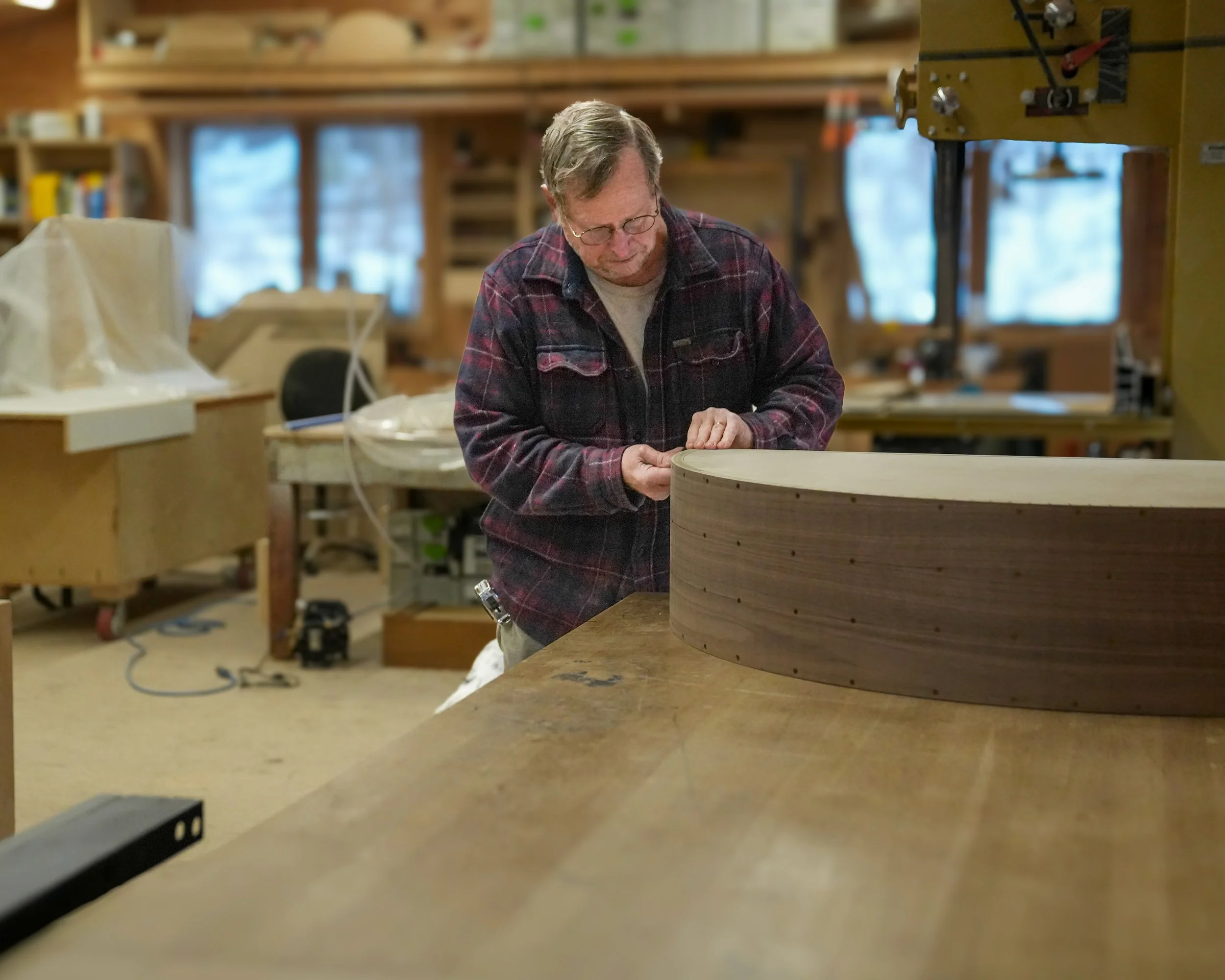 An older man with glasses and a plaid shirt working on a wooden crafts project in a woodworking shop. The shop has shelves, tools, and wood materials, and the man is focused on shaping the wood on a work surface.
