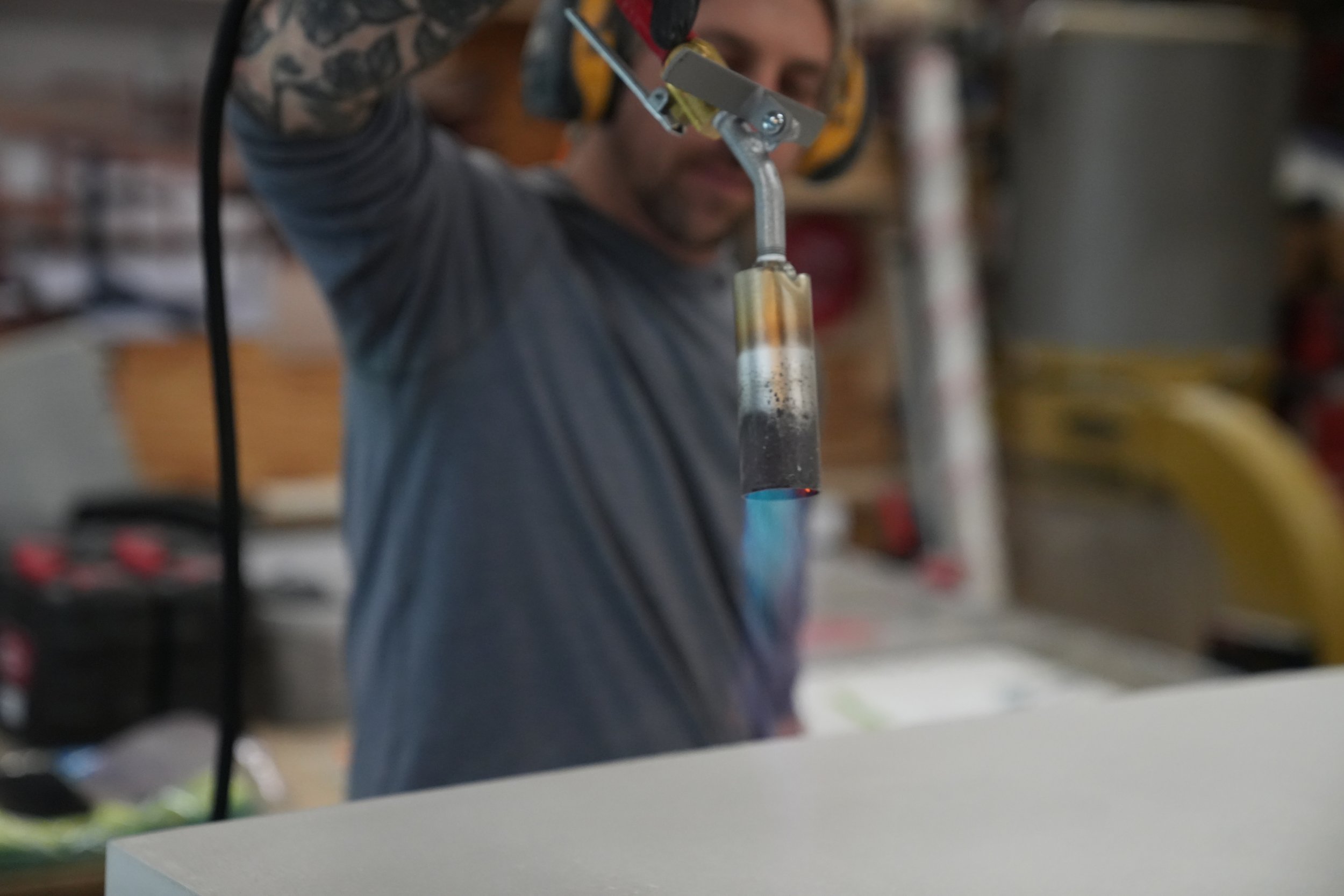 A person with tattoos on their arm is working with a blowtorch, heating a glass tube in a laboratory or workshop setting. The person's face is partially visible, and they are wearing protective ear muffs.