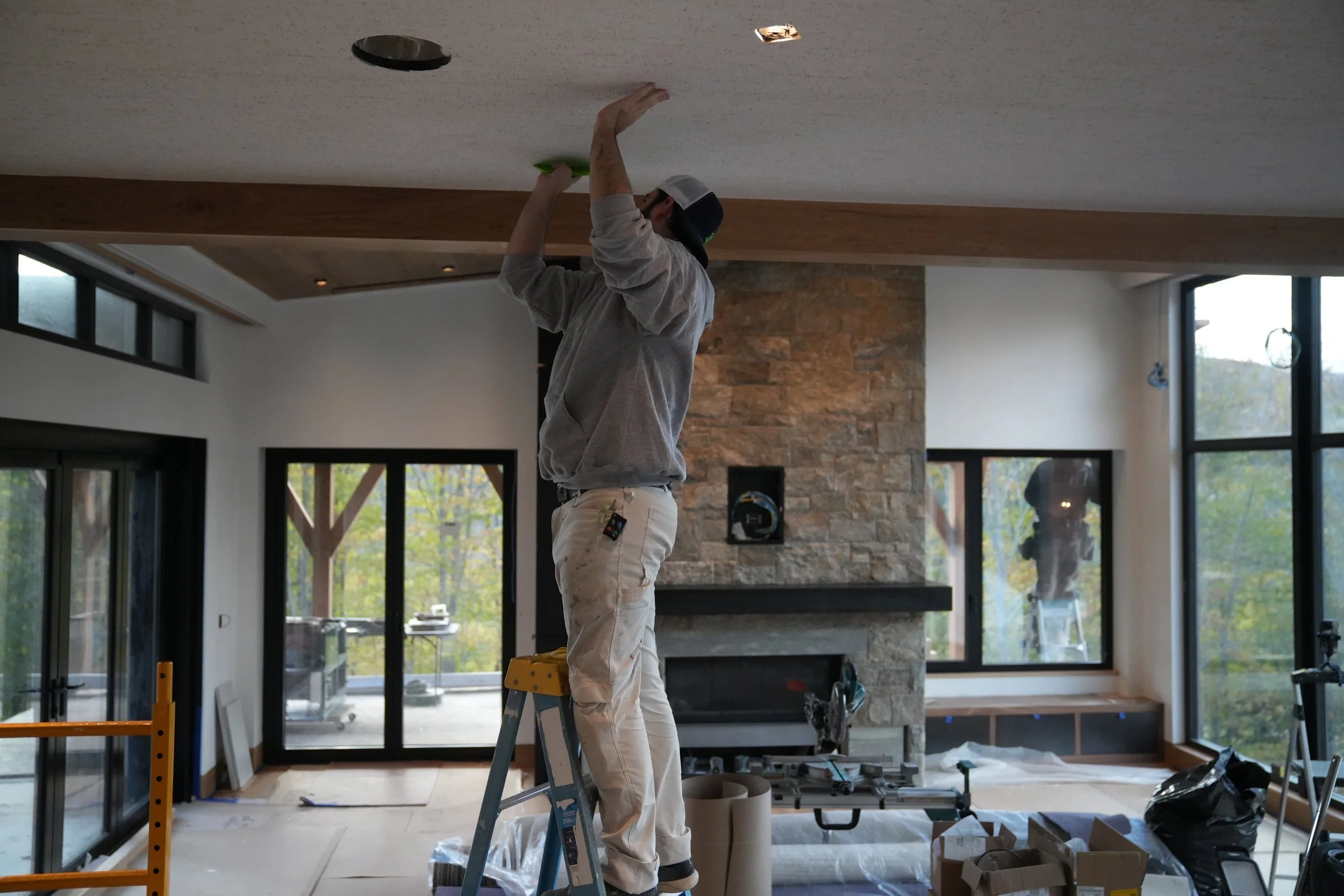 A person wearing a baseball cap and work clothes standing on a ladder, working on a ceiling with exposed lighting fixtures in a modern interior space with large windows and a stone accent wall.