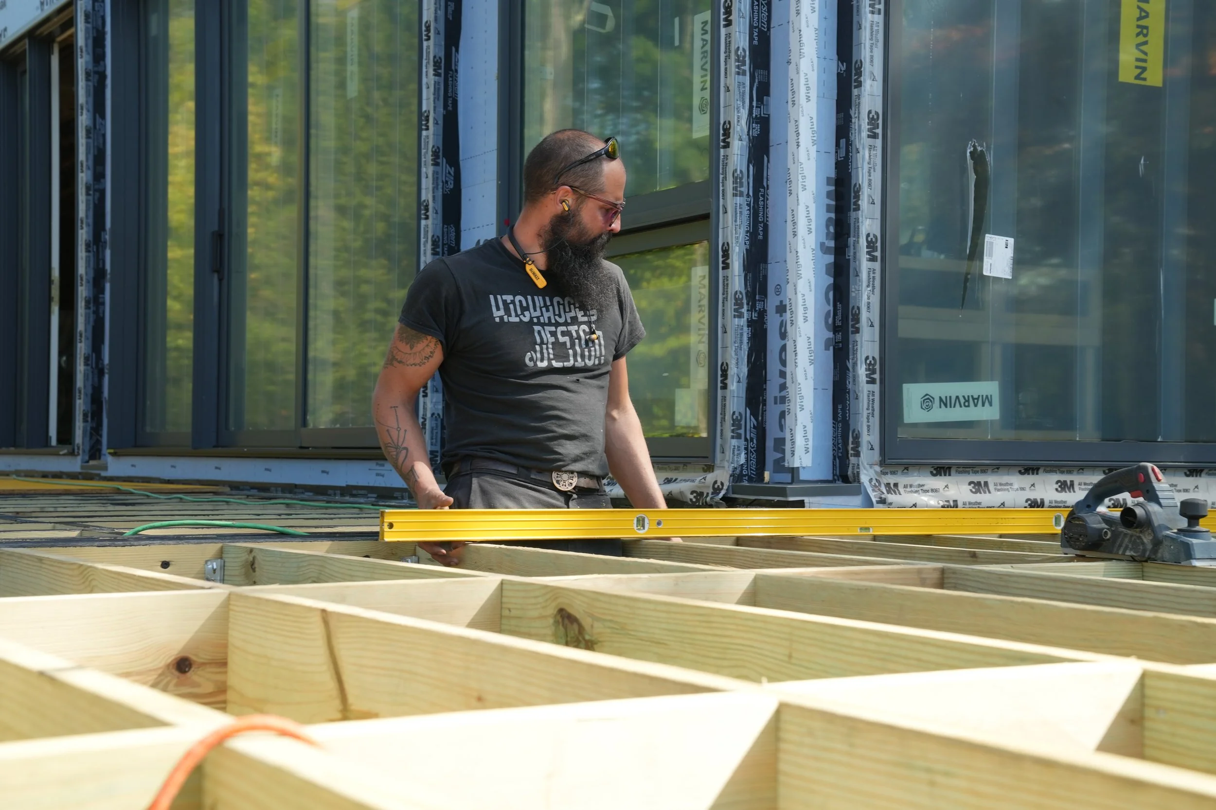 A man with a beard, sunglasses, and tattoos on his arm is working on a construction site, standing amidst unfinished wooden frames with a partially built glass wall behind him.