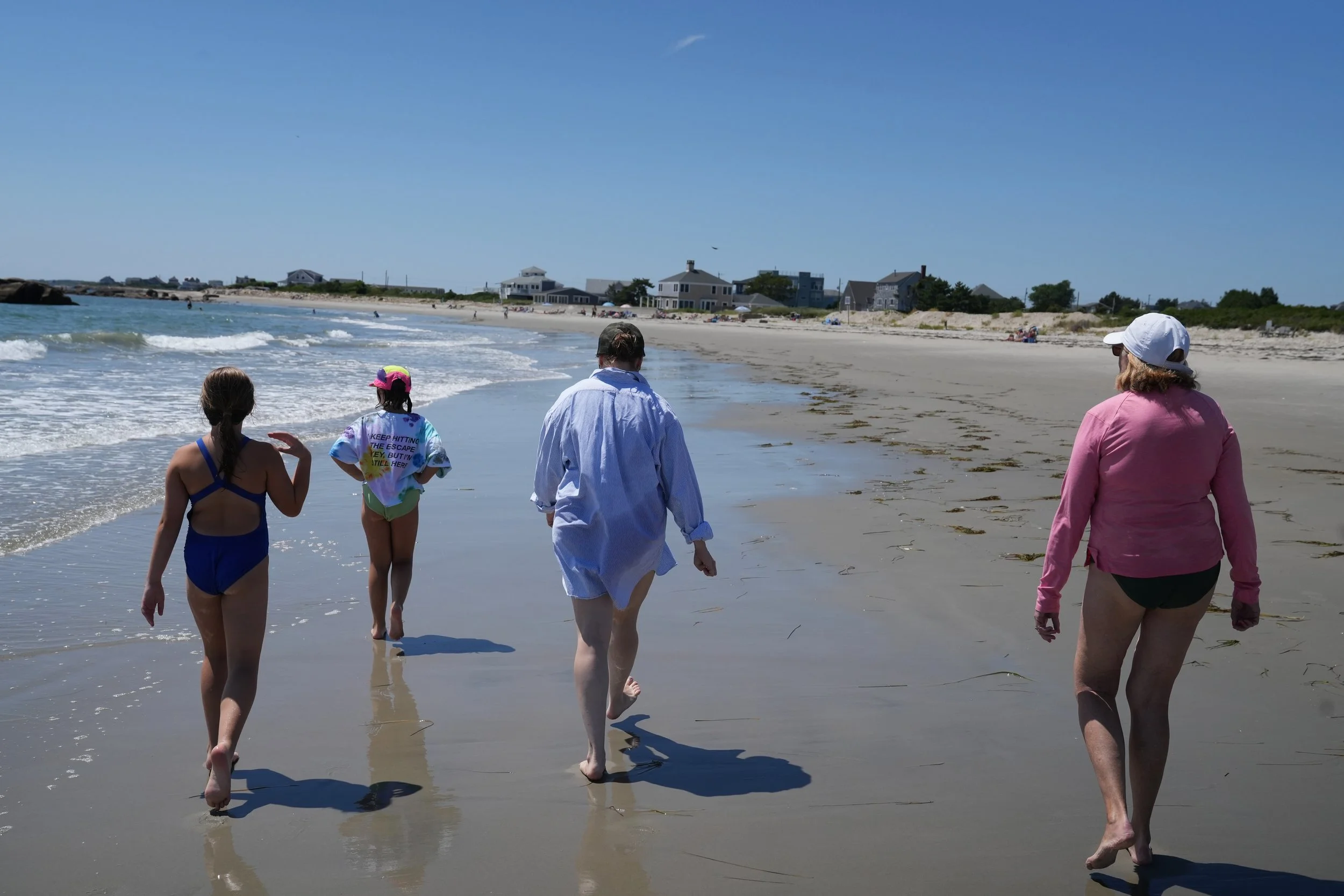 People walking along a sandy beach near the shoreline under a clear blue sky, with houses in the background.