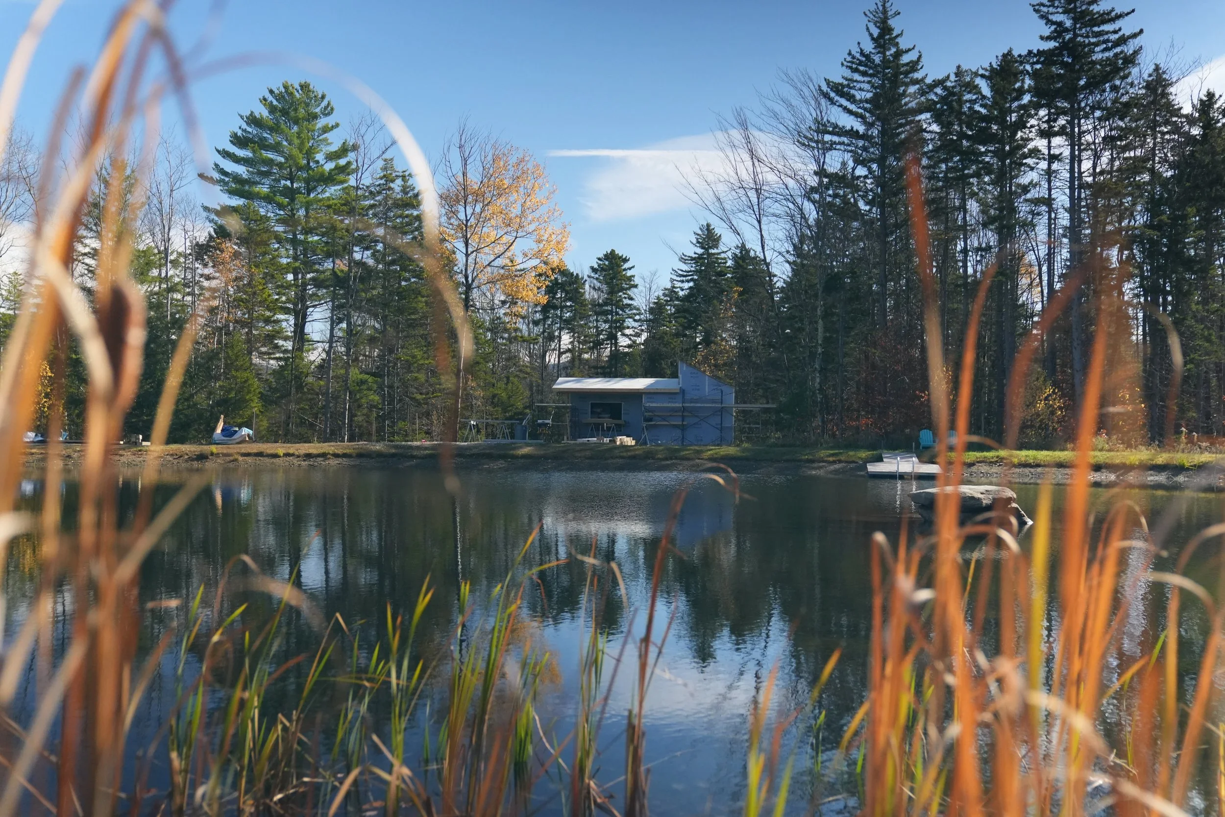 A lake surrounded by tall trees with some changing color, with a small building and boats on the shore, under a clear blue sky.