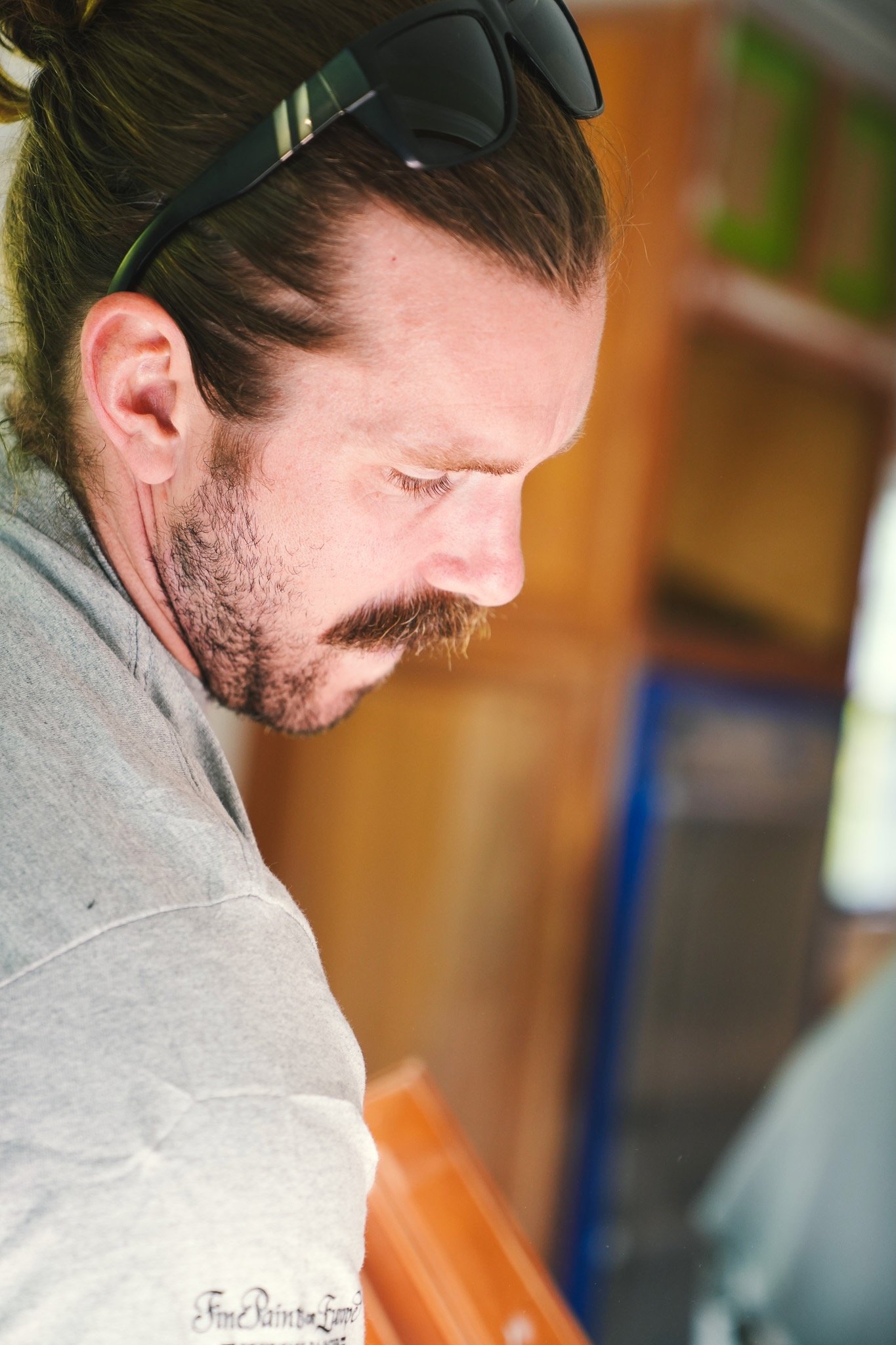 Close-up of a man with a beard and sunglasses resting on his head, looking at a screen. He is wearing a light gray t-shirt.