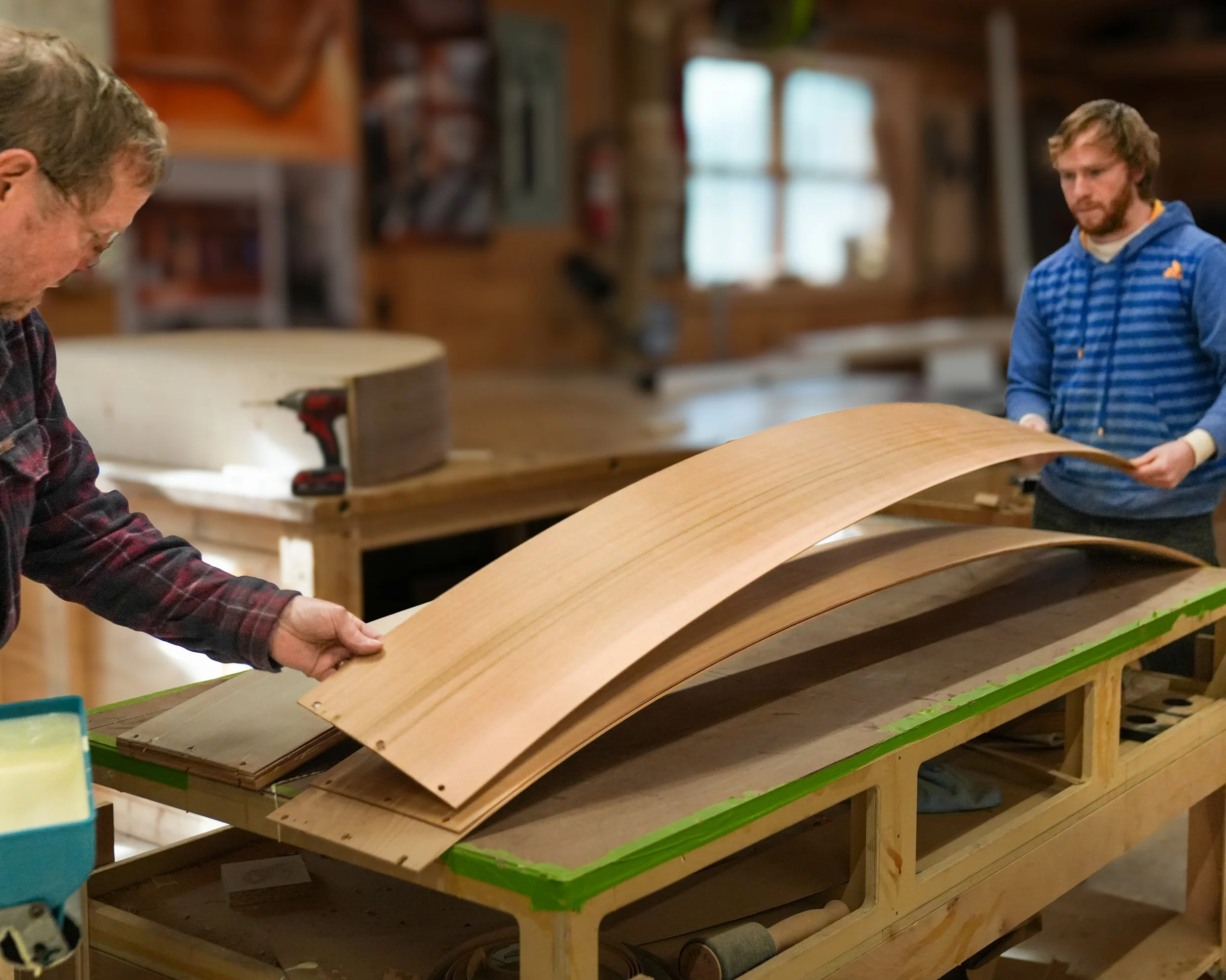 Two men are working on a woodworking project in a workshop. One man on the left is adding a curved wooden piece to a project, while the other man on the right is holding a similar wooden piece. Shelves with supplies and tools are visible in the backg