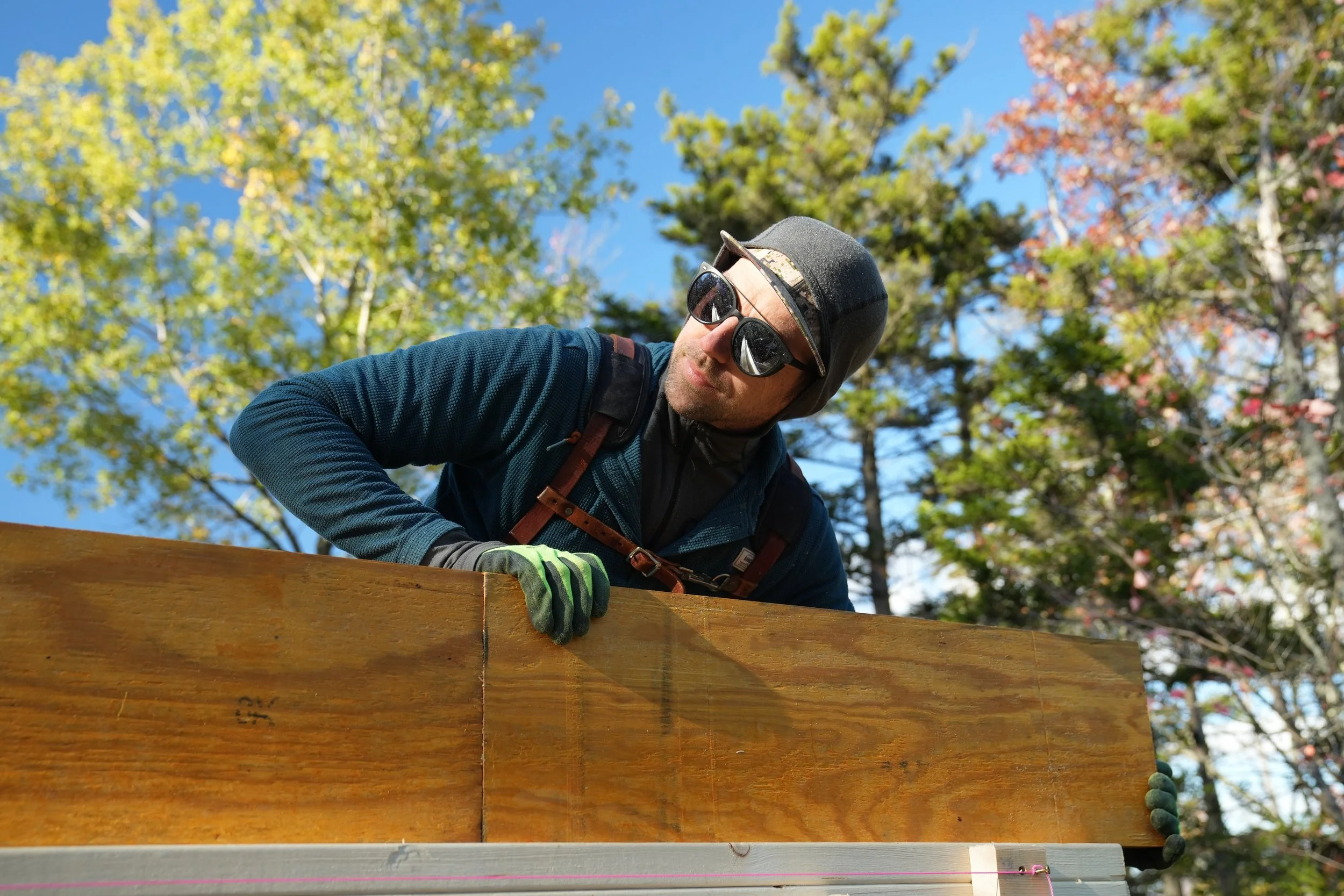 A person wearing a black helmet, sunglasses, and gloves is working outdoors on a wooden structure surrounded by green and autumn-colored trees under a clear blue sky.
