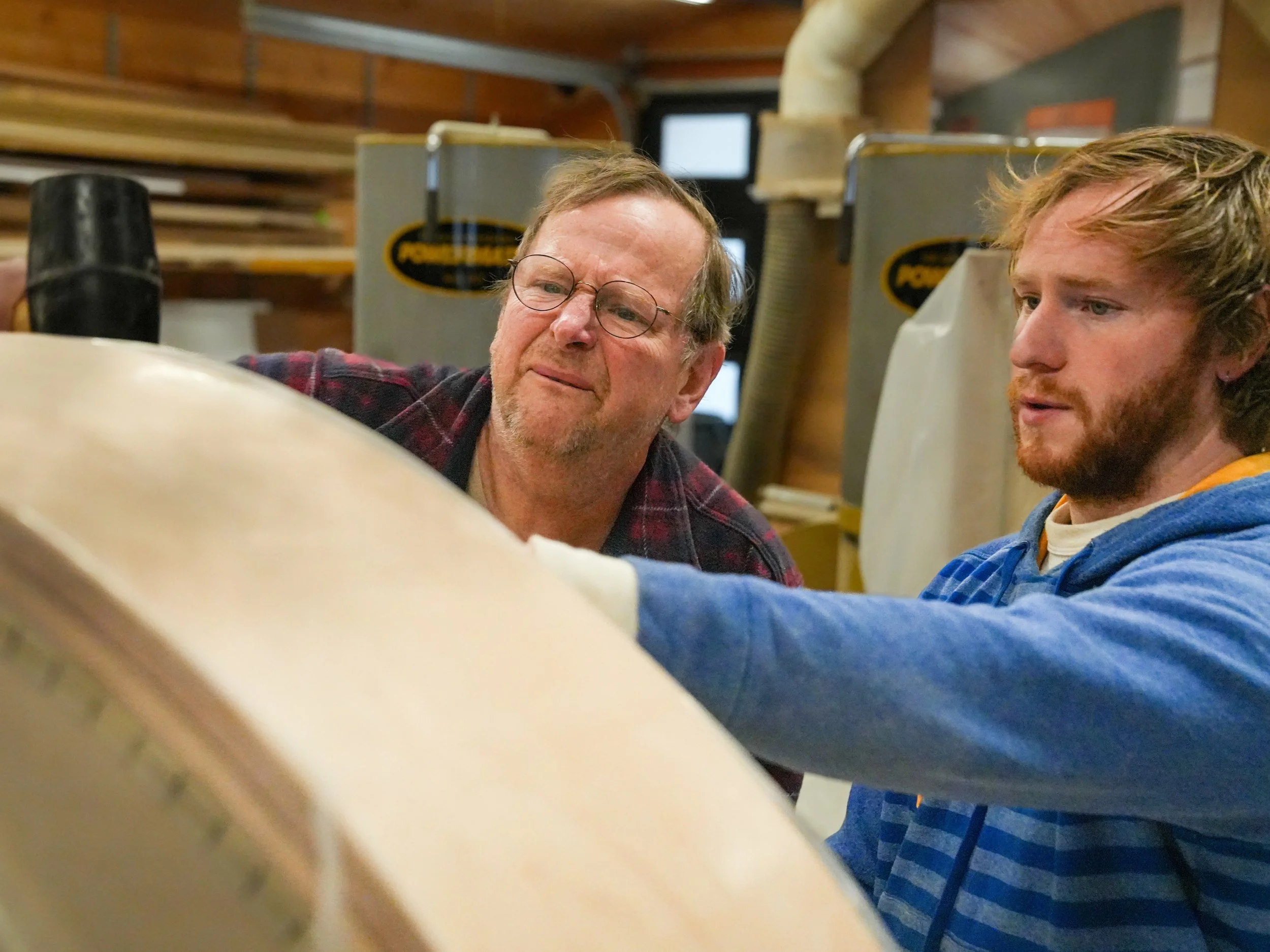 Two men, one older with glasses and a beard, and one younger with a beard and. blond hair, are working together in a woodshop. The older man is looking down at the wood while the younger man is watching.