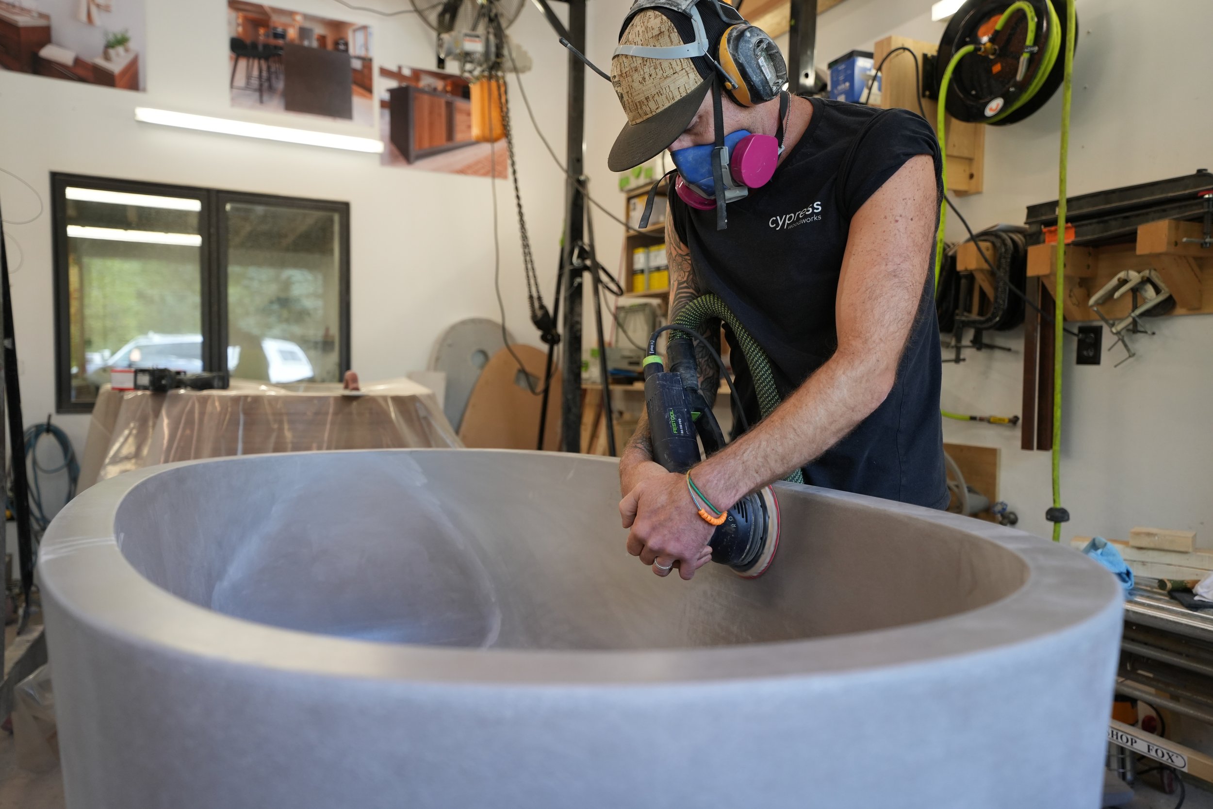 A man wearing a cap, protective ear and face masks, is working on a circular stone or concrete object with a power tool in workshop or studio.