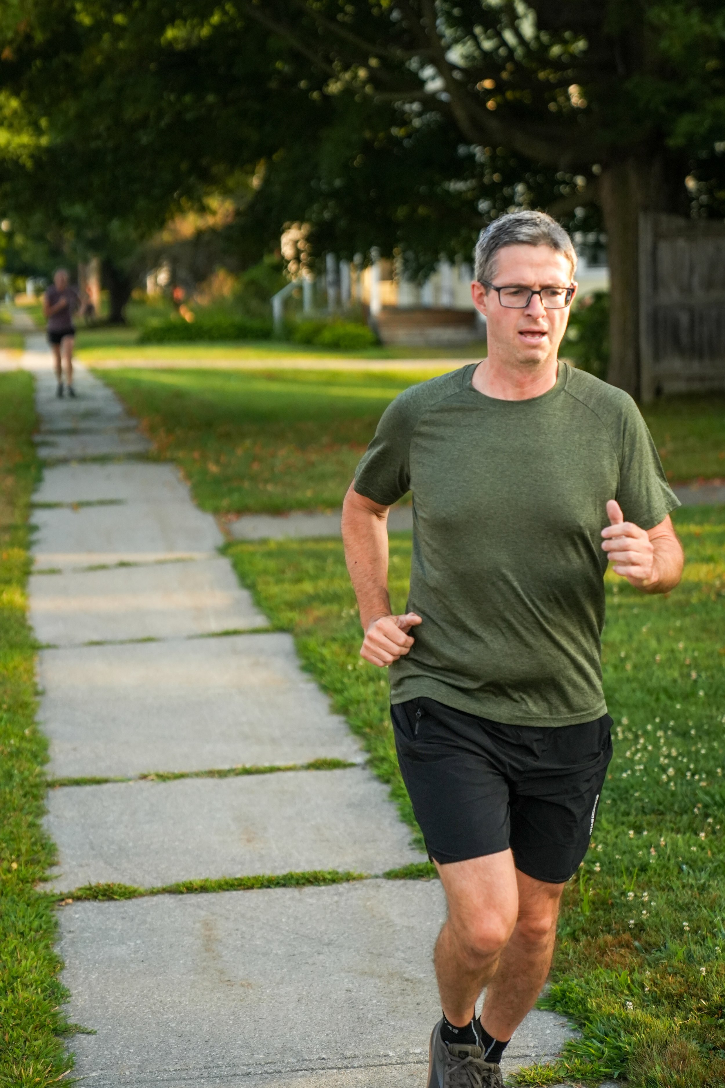 A man jogging on a sidewalk in a neighborhood with green trees and grass, wearing a green t-shirt, black shorts, and running shoes.
