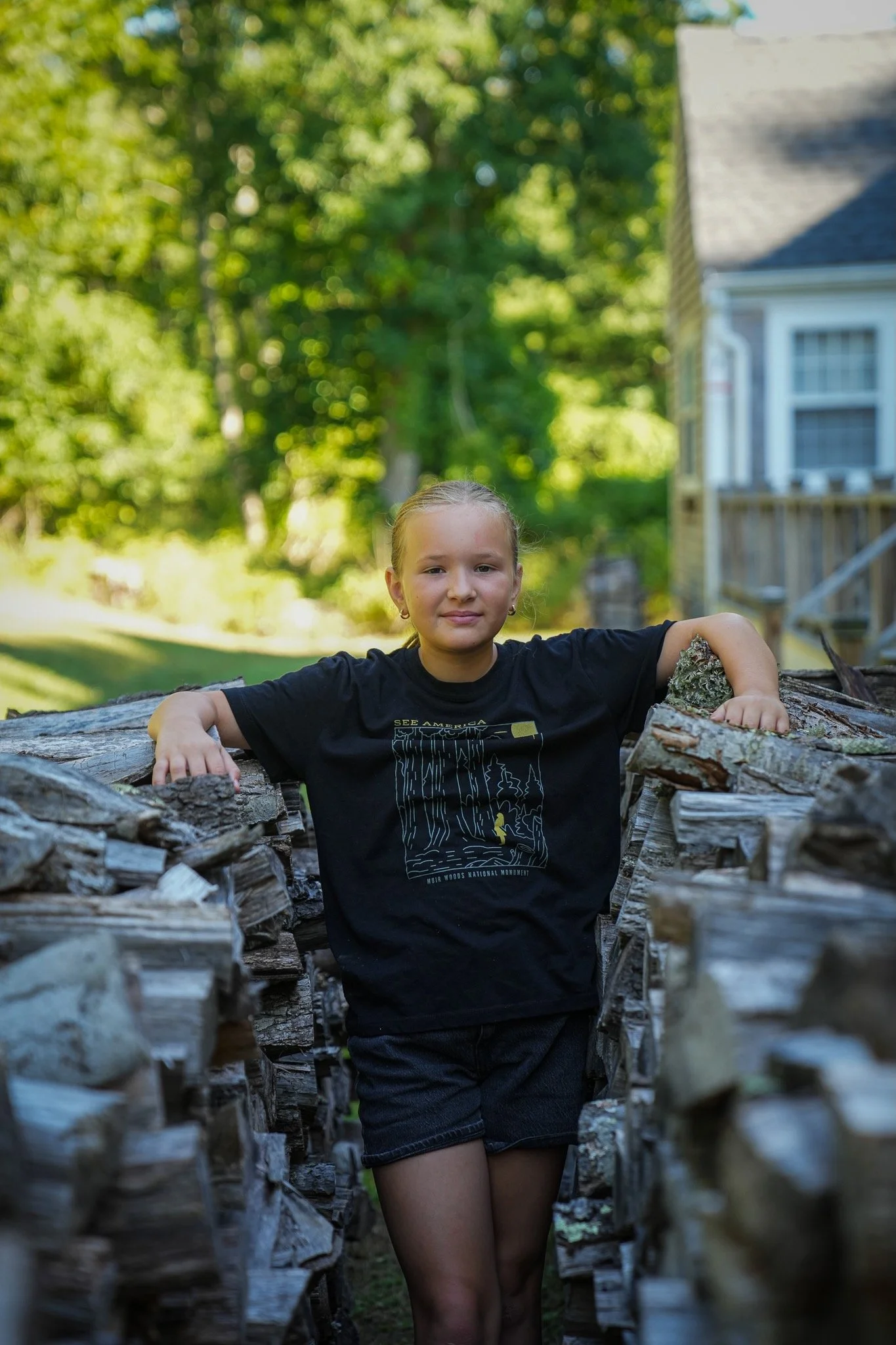 A young girl with Blonde hair in a bun, wearing a black T-shirt with a graphic design on the front, standing in a narrow pathway between stacked rows of cut wood logs outdoors. Green trees and a house with a porch are visible in the background.