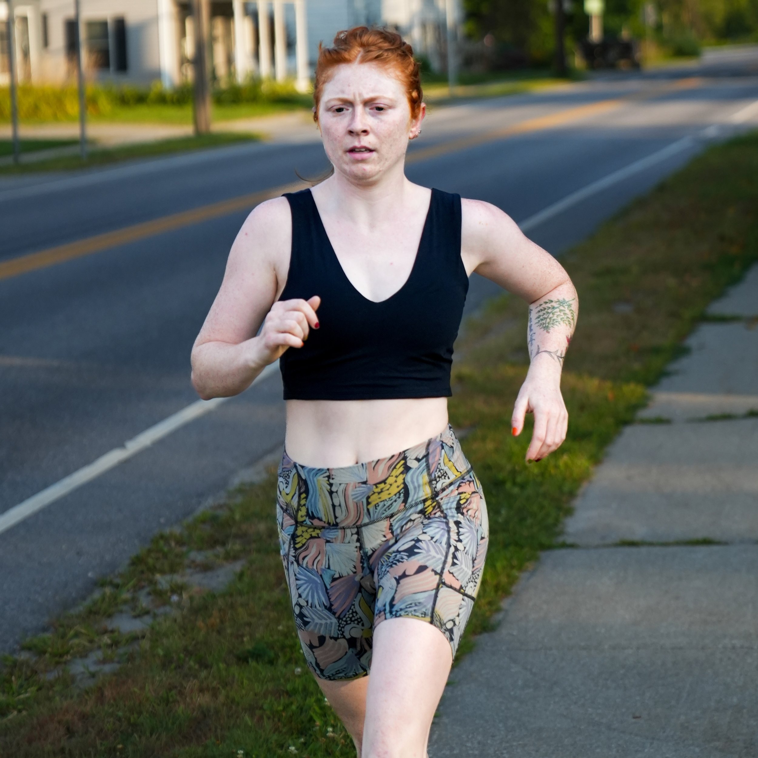 A woman jogging outdoors on a suburban street with trees and houses in the background, wearing a black sports top and patterned shorts.