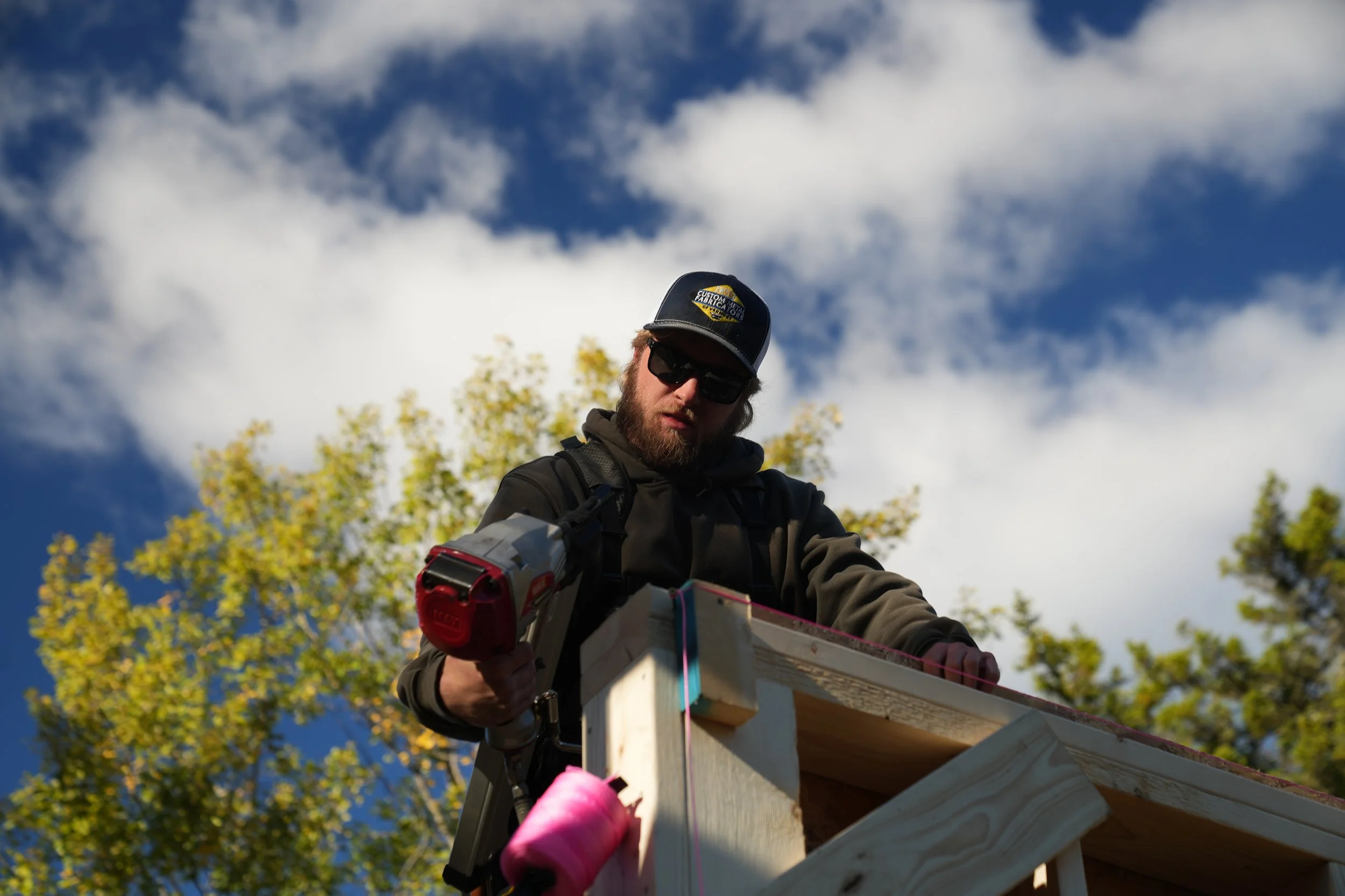 Man wearing sunglasses, a dark cap with a yellow logo, and a dark jacket working on a wooden construction project outdoors, with trees and a partly cloudy sky in the background.