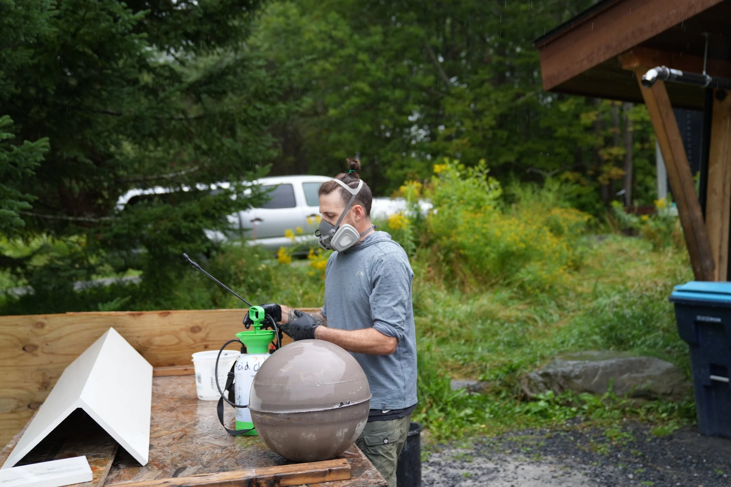 A person wearing a mask and gloves is working outdoors with a large, round ceramic object, possibly for a kiln, on a wooden table under a roofed area. There are containers and equipment around. The background shows green trees, yellow flowers, and a 