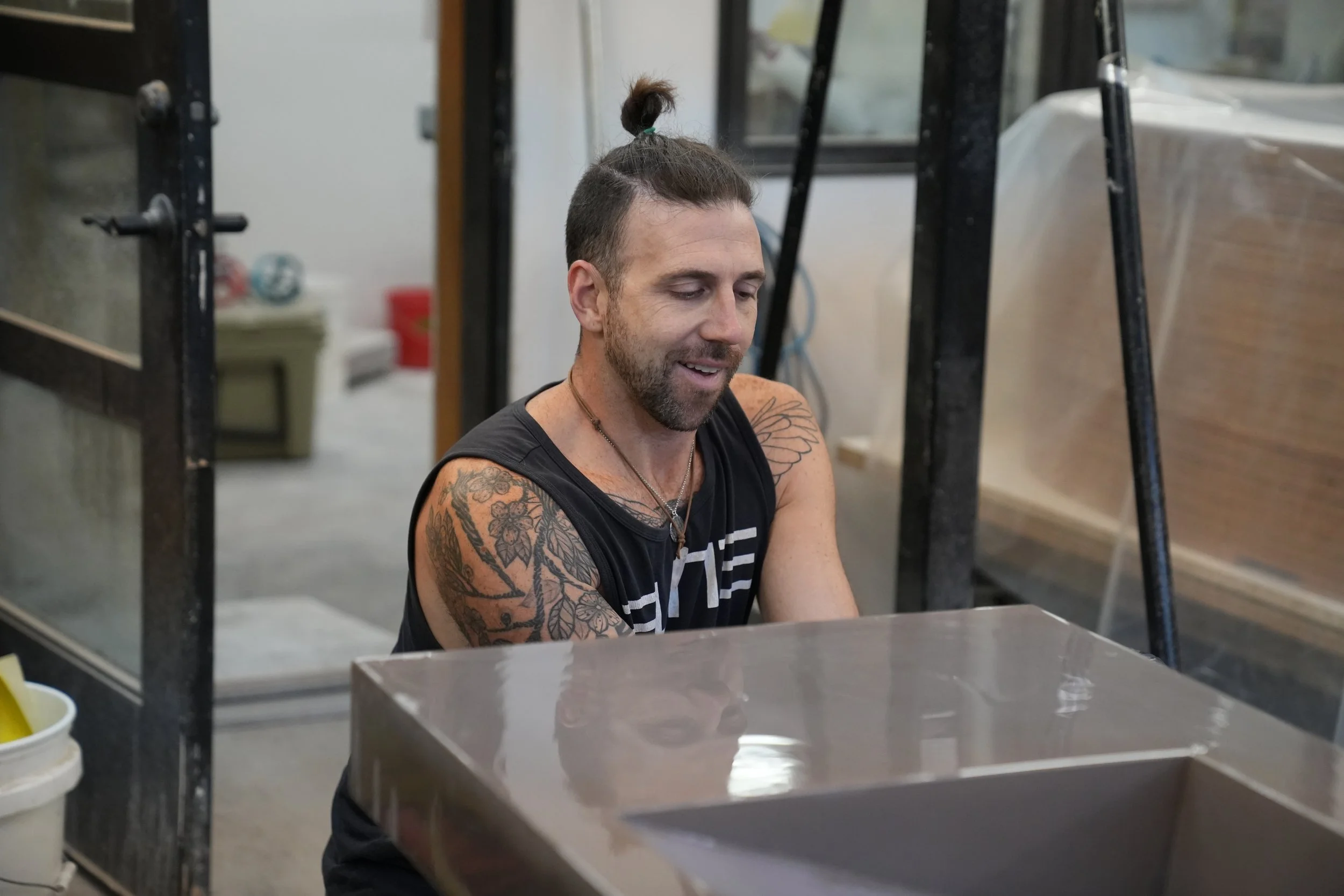 A man with tattoos on his right arm, dark hair in a top knot, and a beard is sitting at a counter in a workshop or garage. He is smiling and looking down. There is a black metal shelving unit and a plastic container in the foreground, with a red cont
